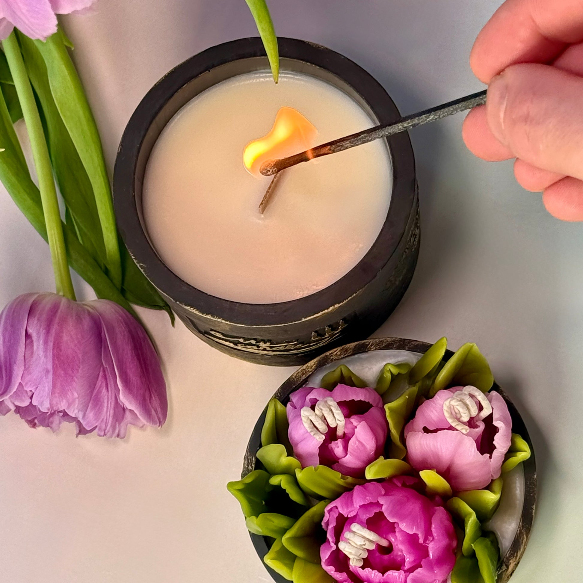 Lighting a wooden wick crackling candle inside a handcrafted black Jesmonite vessel, next to a removable lid decorated with realistic purple beeswax tulip flowers.