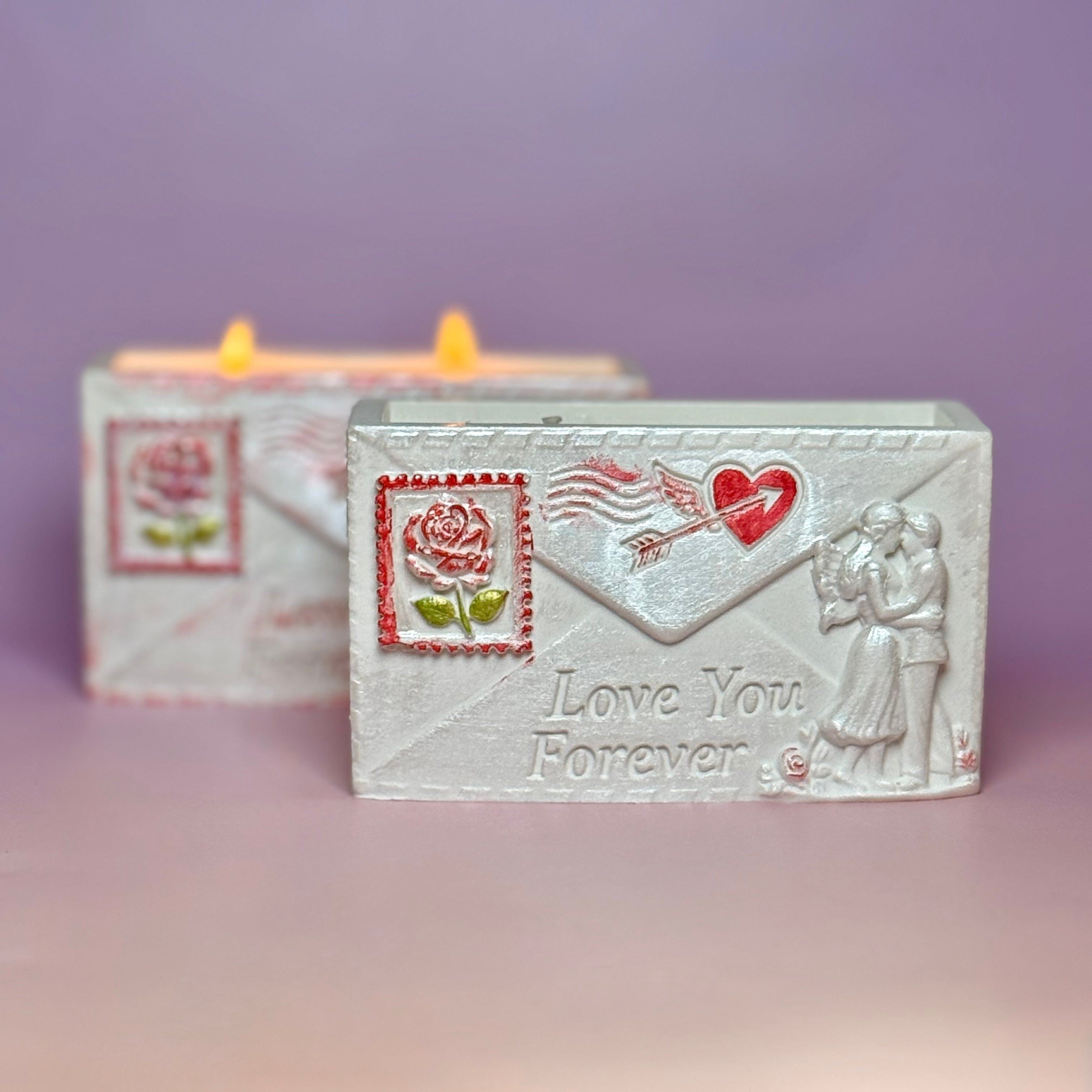 A pearl-white Jesmonite envelope candle holder with the inscription Love You Forever, a hand-painted red rose stamp, and a lit double-wick soy candle burning in the background.