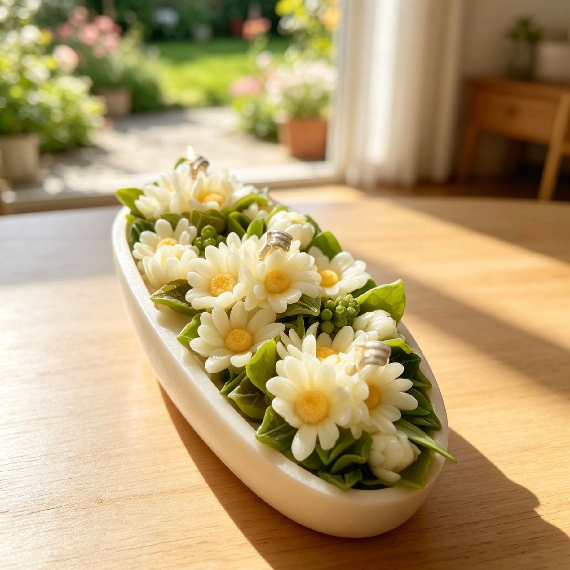 A beautiful daisy bouquet candle arrangement in an oval white Jesmonite bowl, placed on a sunlit wooden table with a blurred garden view background.