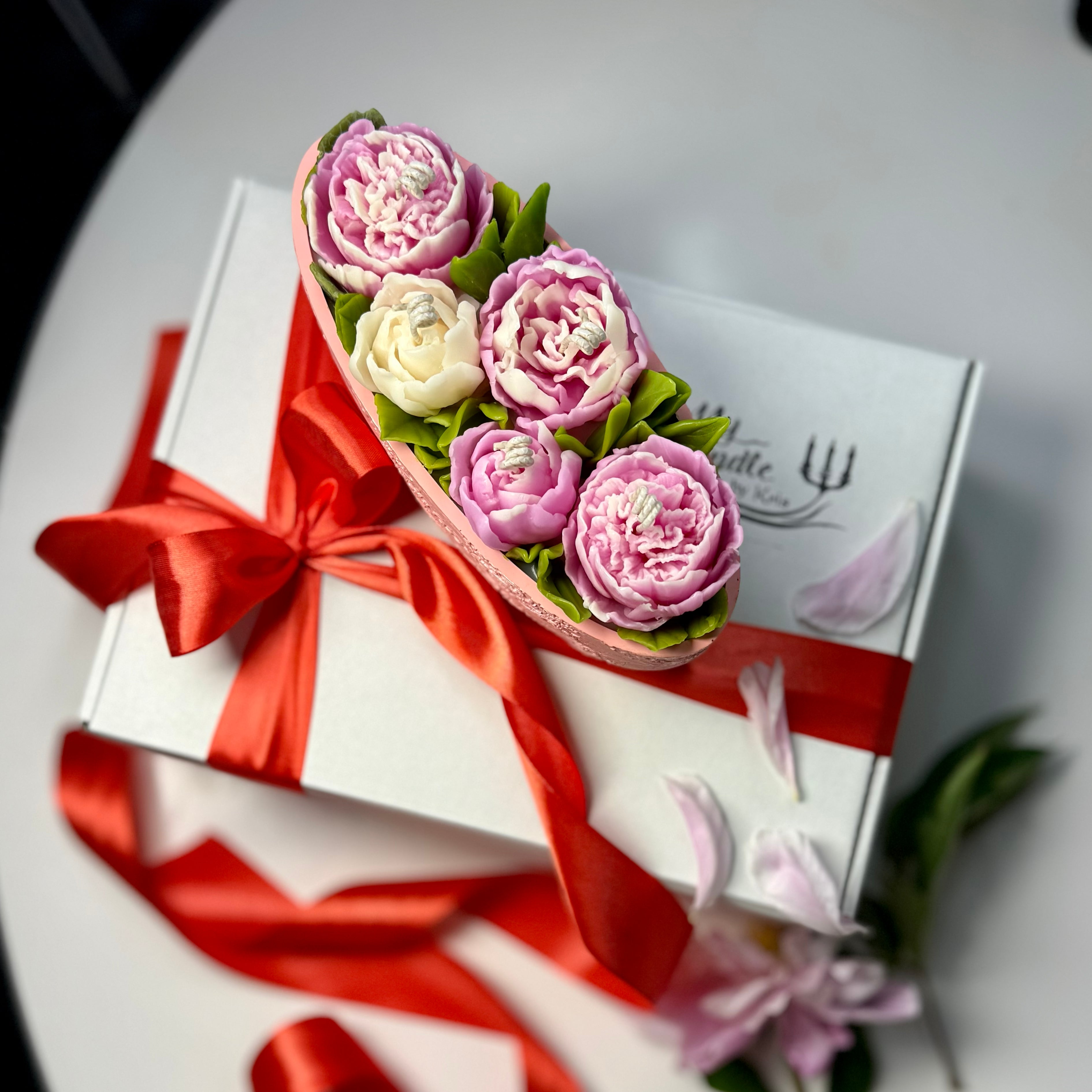 Overhead view of a pink Jesmonite floral candle featuring hand-sculpted pink and white beeswax peonies on top of a white gift box tied with a satin red ribbon.
