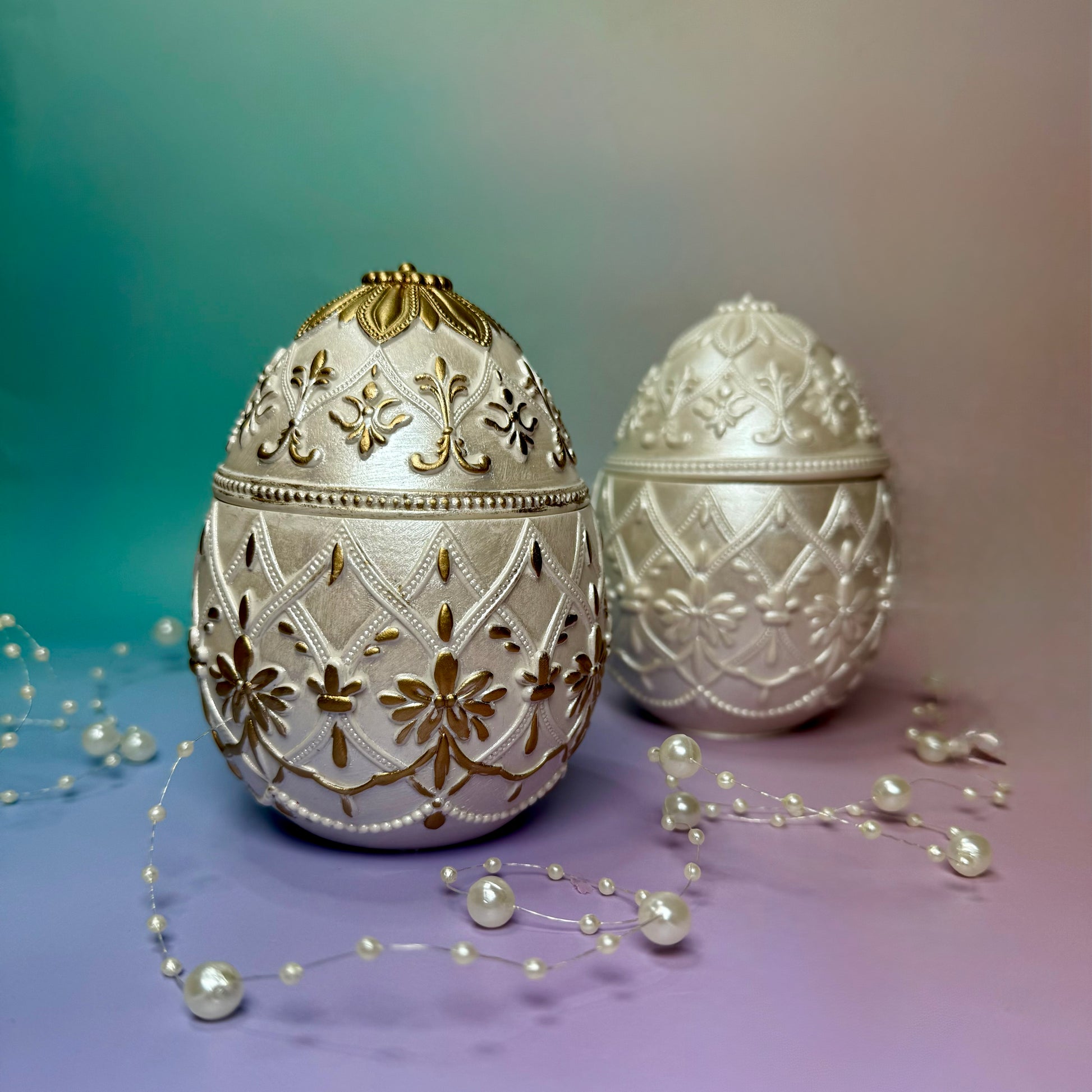 Two luxury ornate Easter egg candles cast in Jesmonite, one hand-painted with gold detailing and one in pearlescent classic white, styled with pearls.
