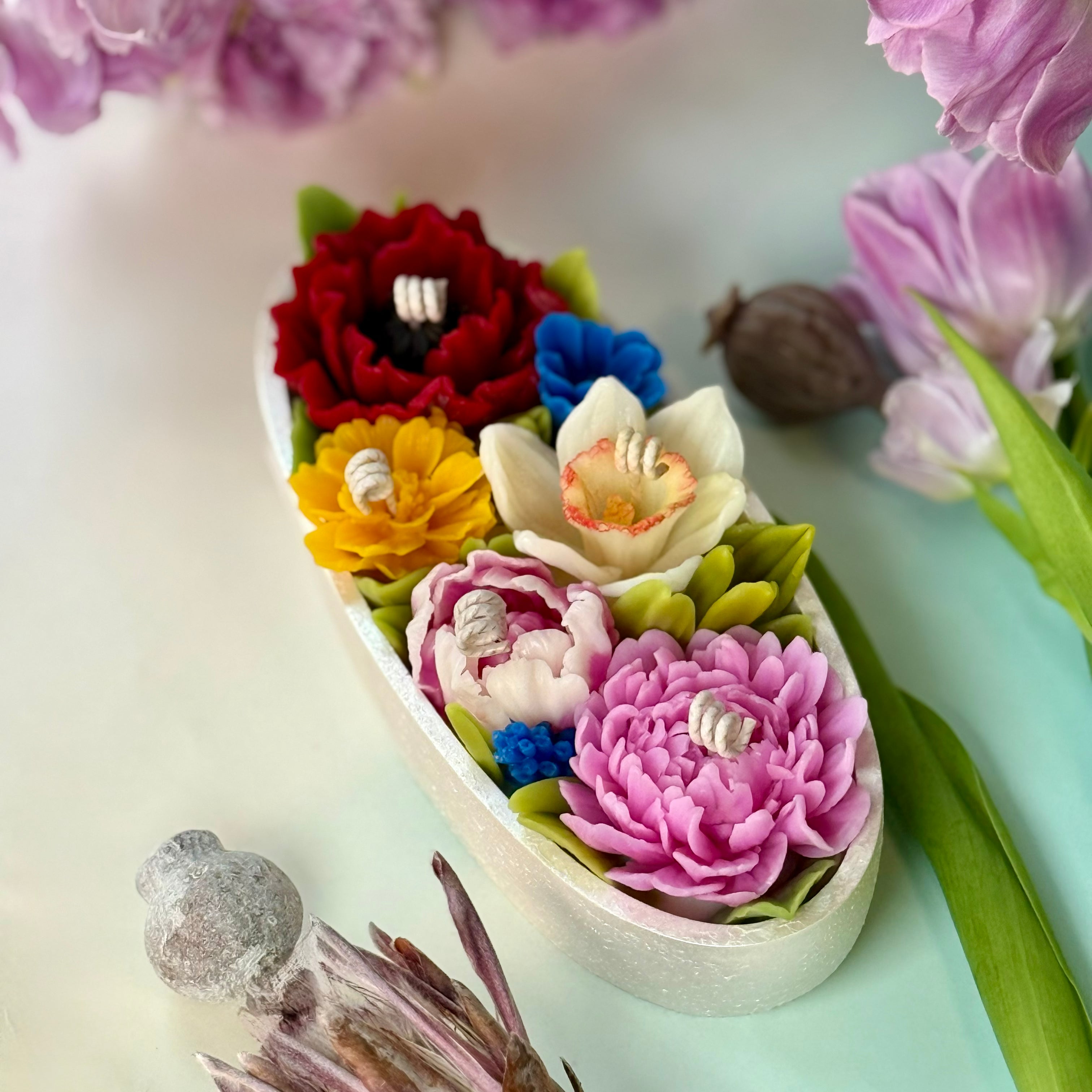 Elegant full-length view of a luxury oval botanical candle boat featuring artisan multi-coloured spring flowers and green leaves on a pastel background with gift box.