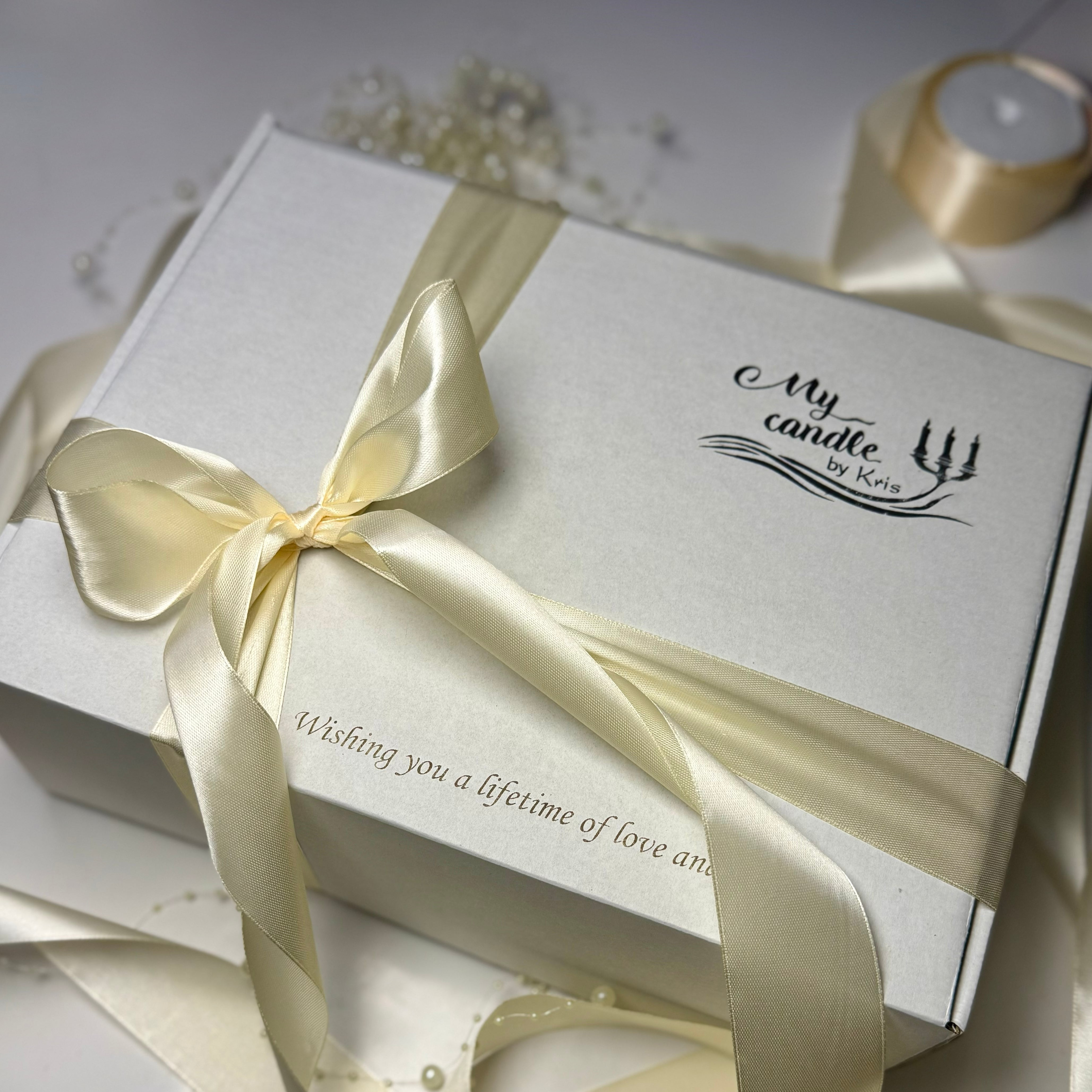 An angled shot of a white premium gift presentation box from My Candle by Kris, wrapped with a ivory satin ribbon and showcasing fine laser engraving that says Wishing you a lifetime of love and.