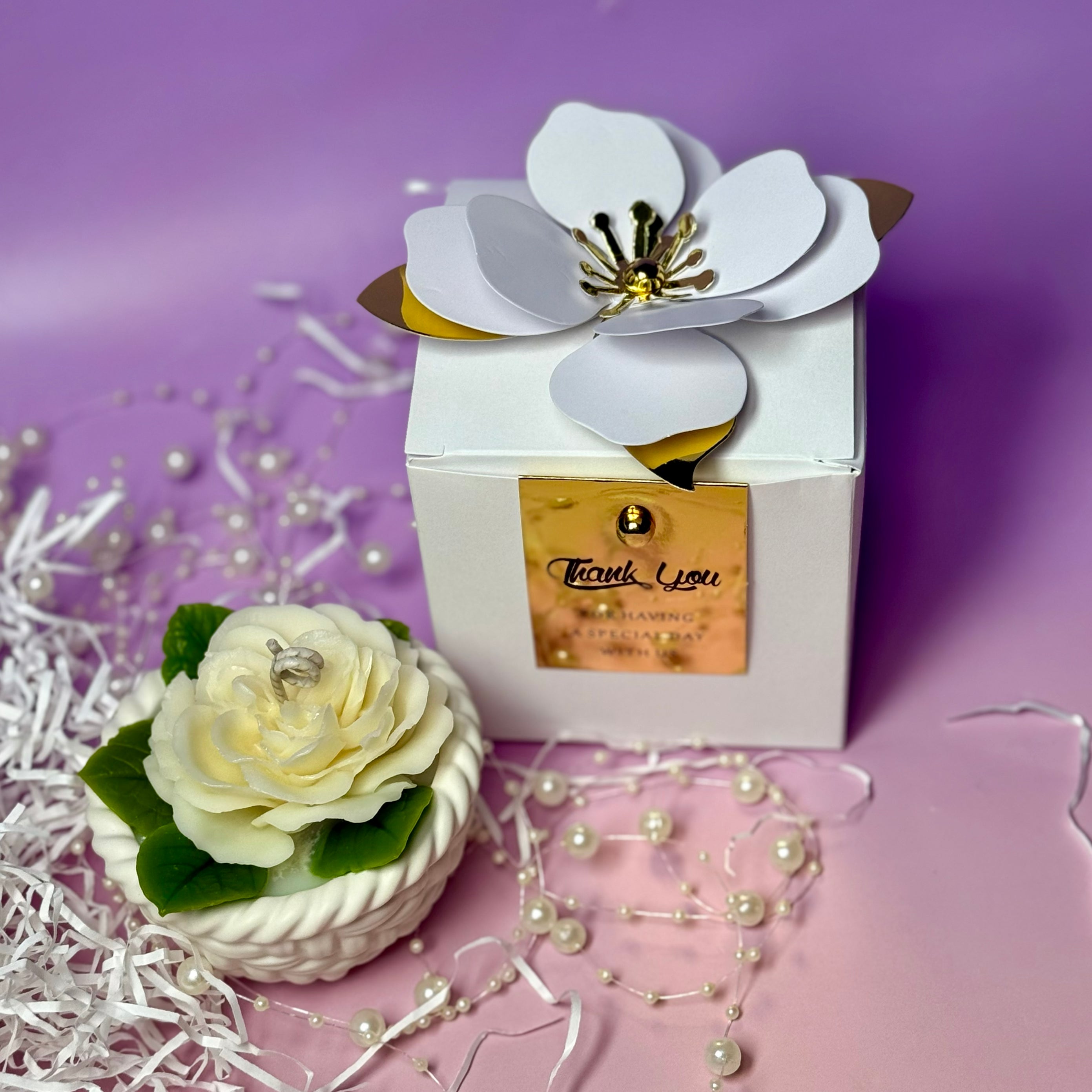 A styled promotional photo showing the cream Abraham Darby rose basket candle sitting on crinkle packaging paper next to a white gift box with a gold foil label.