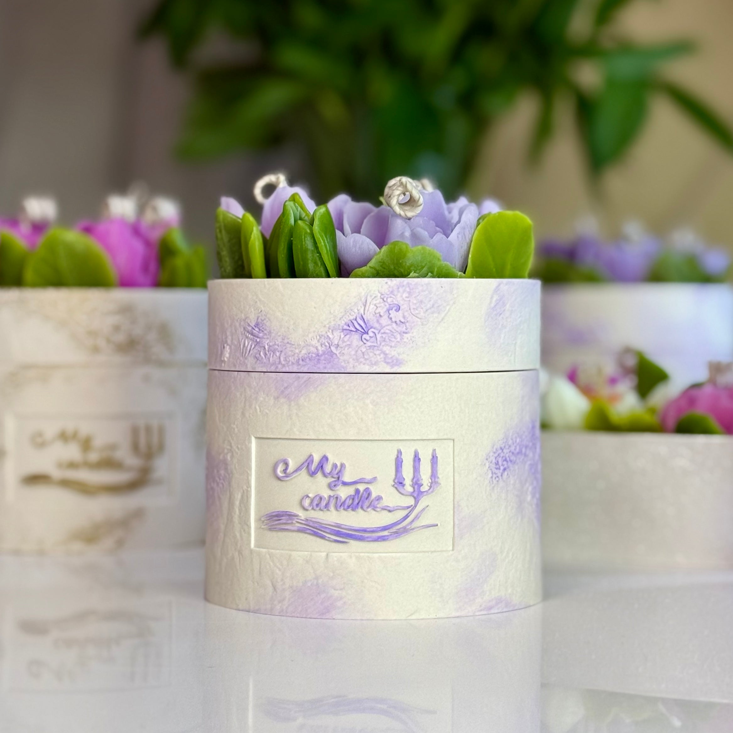 Front view of an artisan round floral candle in a white stone-textured Jesmonite pot with purple accents, highlighting realistic purple beeswax flowers with gift box.