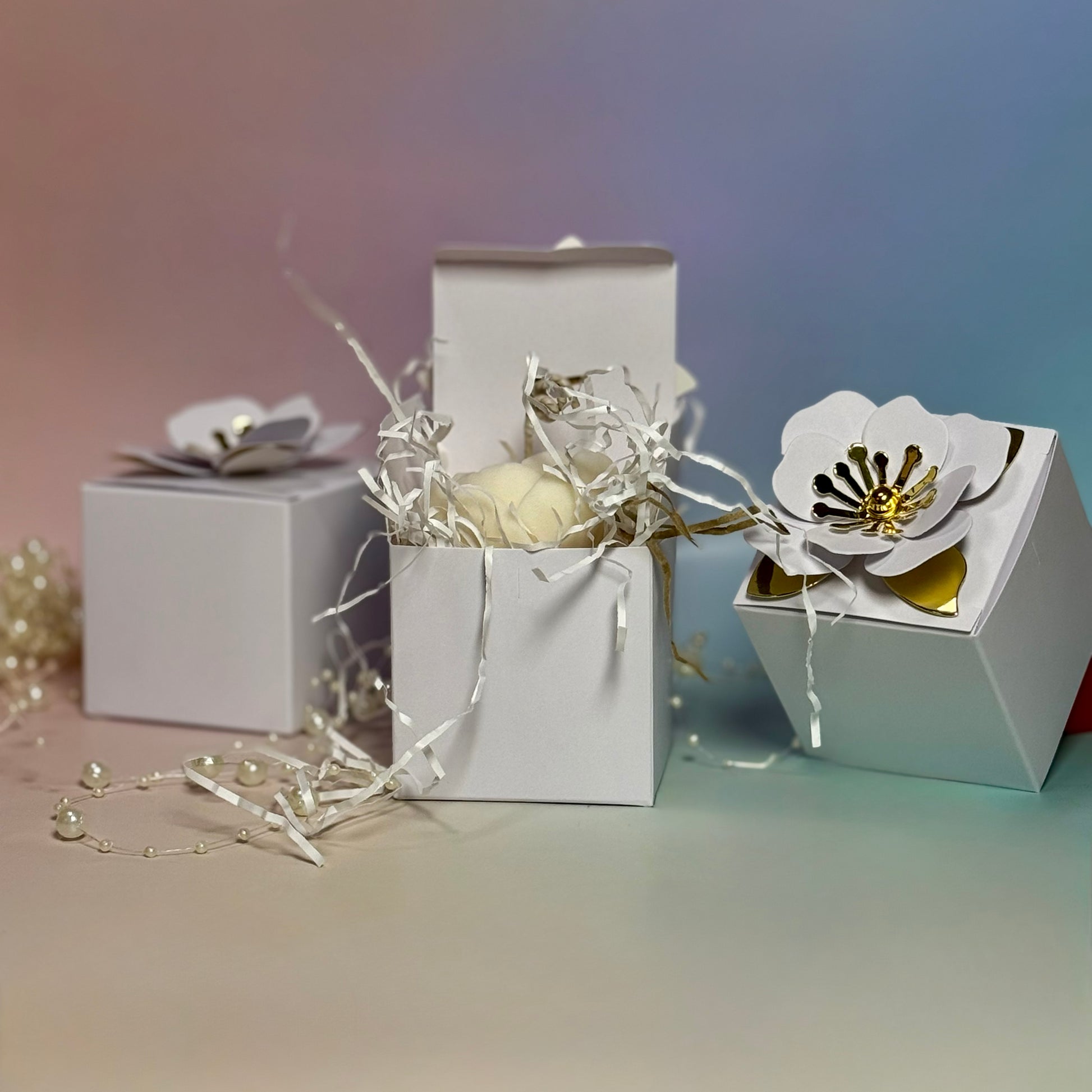 Three white wedding favour gift boxes, with the central box opened to reveal a creamy white soy wax peony candle nestled inside protective white crinkle paper shred.