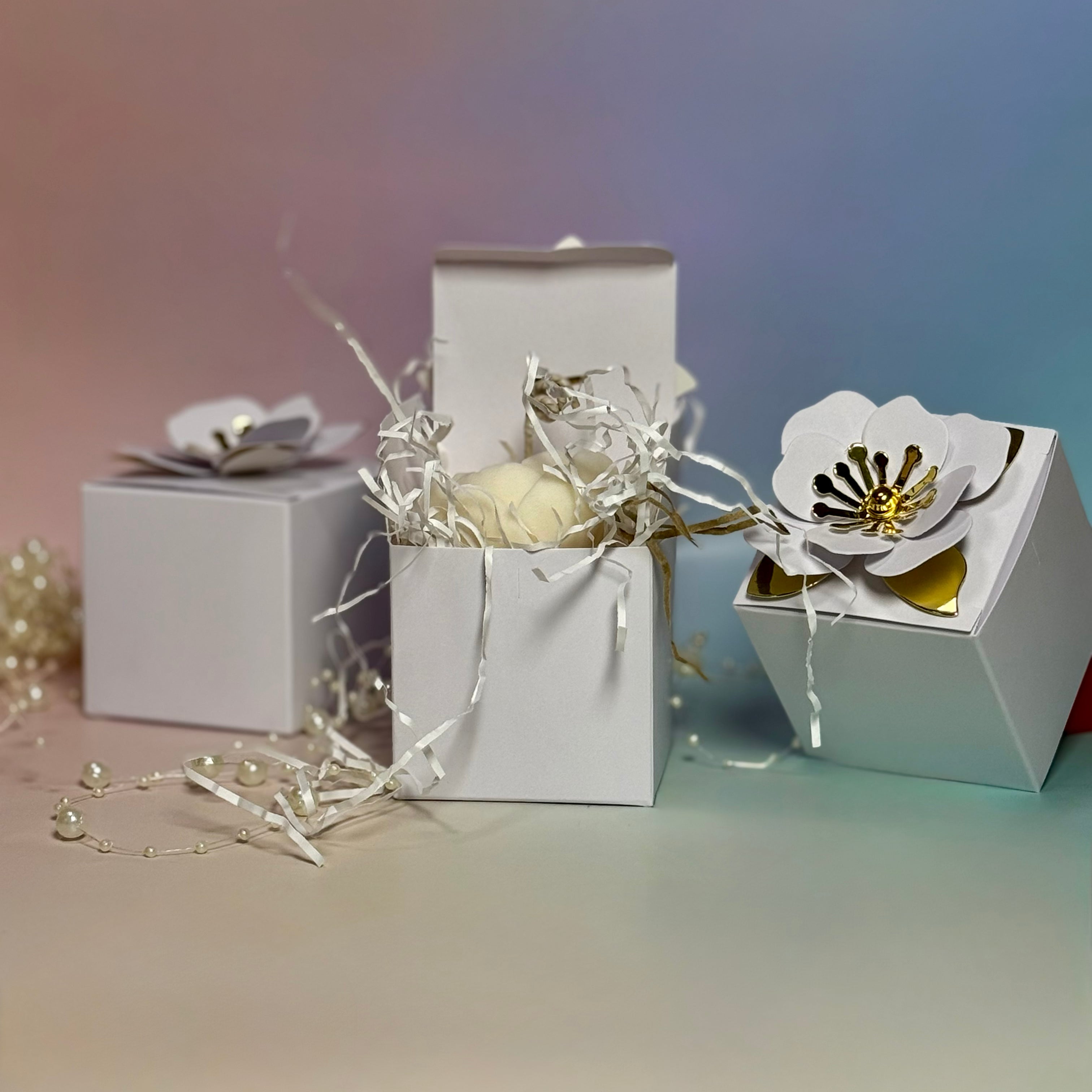 Three white wedding favour gift boxes, with the central box opened to reveal a creamy white soy wax peony candle nestled inside protective white crinkle paper shred.