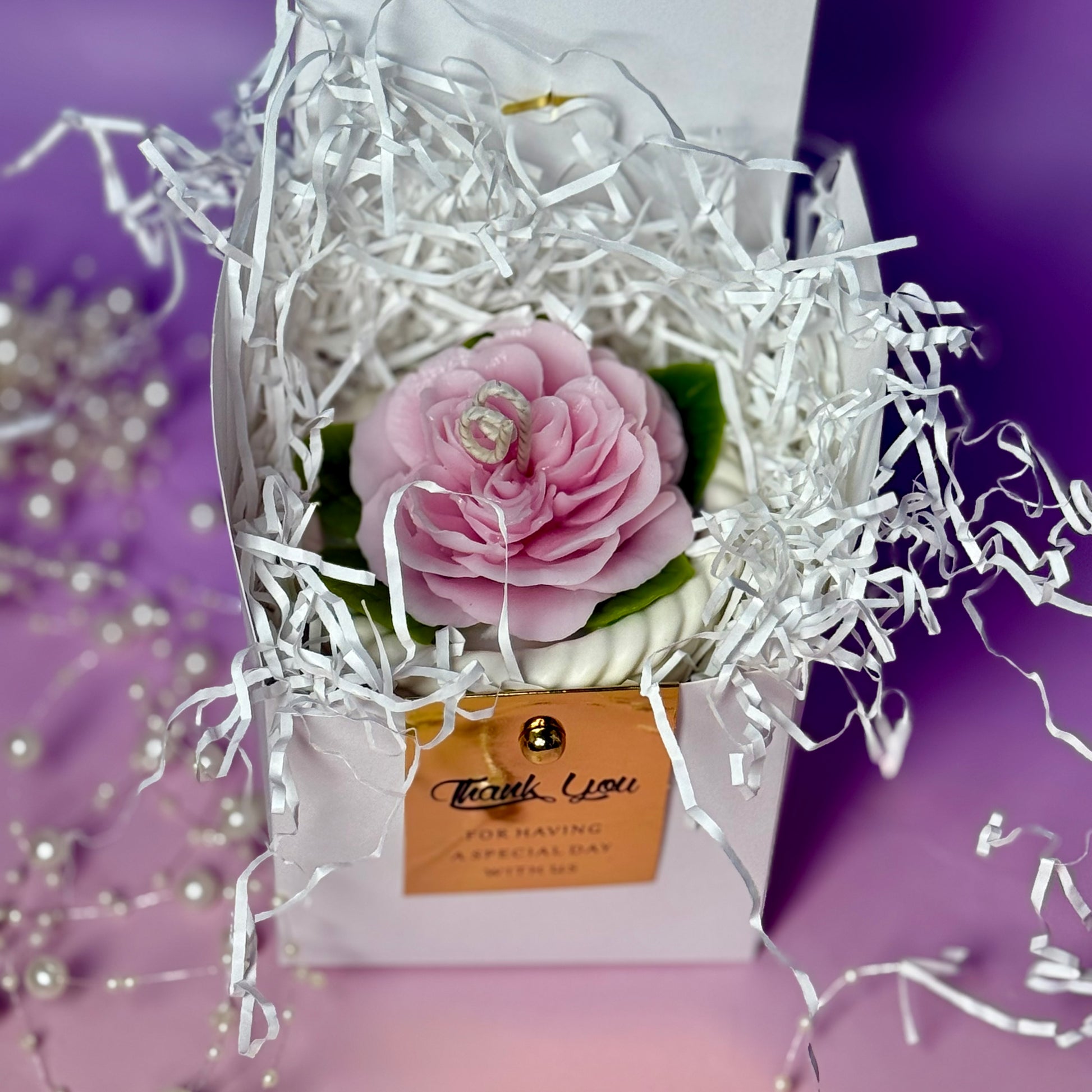 A top-down view into an opened luxury gift box packed with crinkle shred paper, protecting a beautiful pink beeswax rose flower basket candle.
