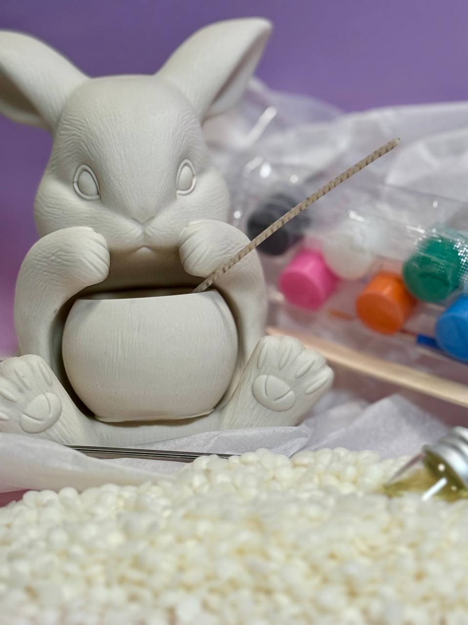 Close-up of a make your own candle kit featuring a white hand-cast Jesmonite bunny holding an integrated candle vessel, set on top of natural soy wax flakes with paint sets in the background.
