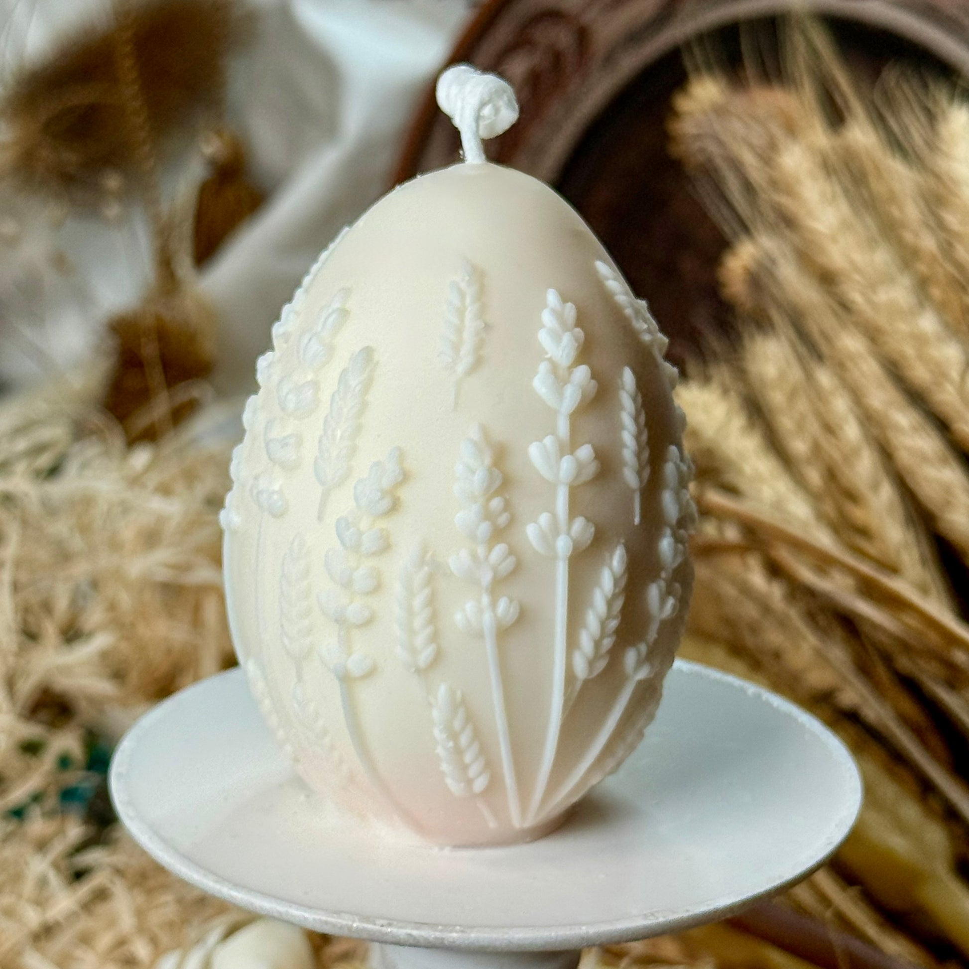 Side view of the unpainted natural ivory wheat stalk and lavender relief egg candle resting on a small white pedestal dish with dried grasses blurred in the background.
