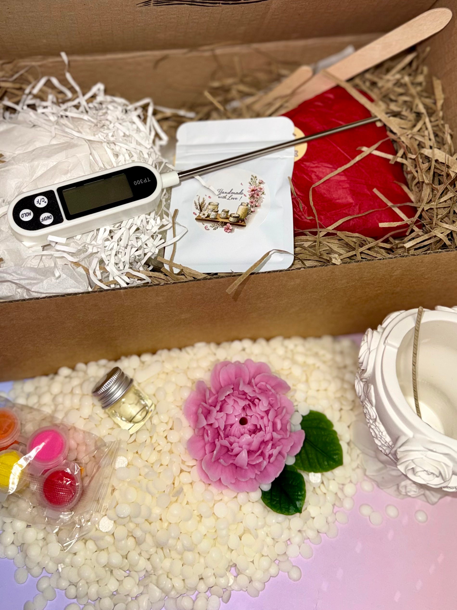 Overhead view of the luxury DIY candle kit contents, displaying the digital thermometer probe, stirring sticks, raw materials, acrylic paints, and a detailed pink beeswax floral topper on wax pellets.