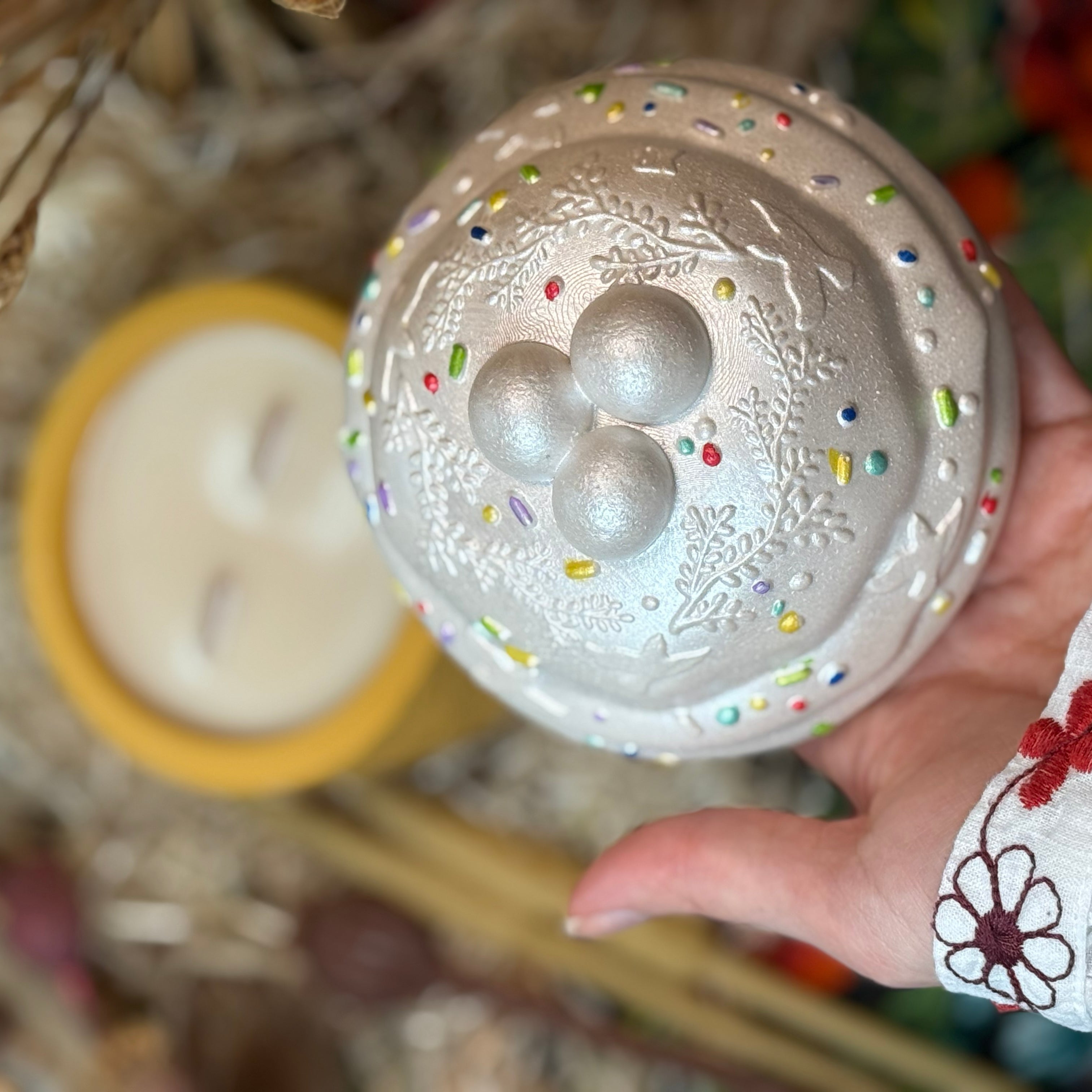 A top-down view showing a hand lifting the decorative lid of the Paska candle to reveal the natural soy wax inside with a double wooden wick, set against a rustic background.
