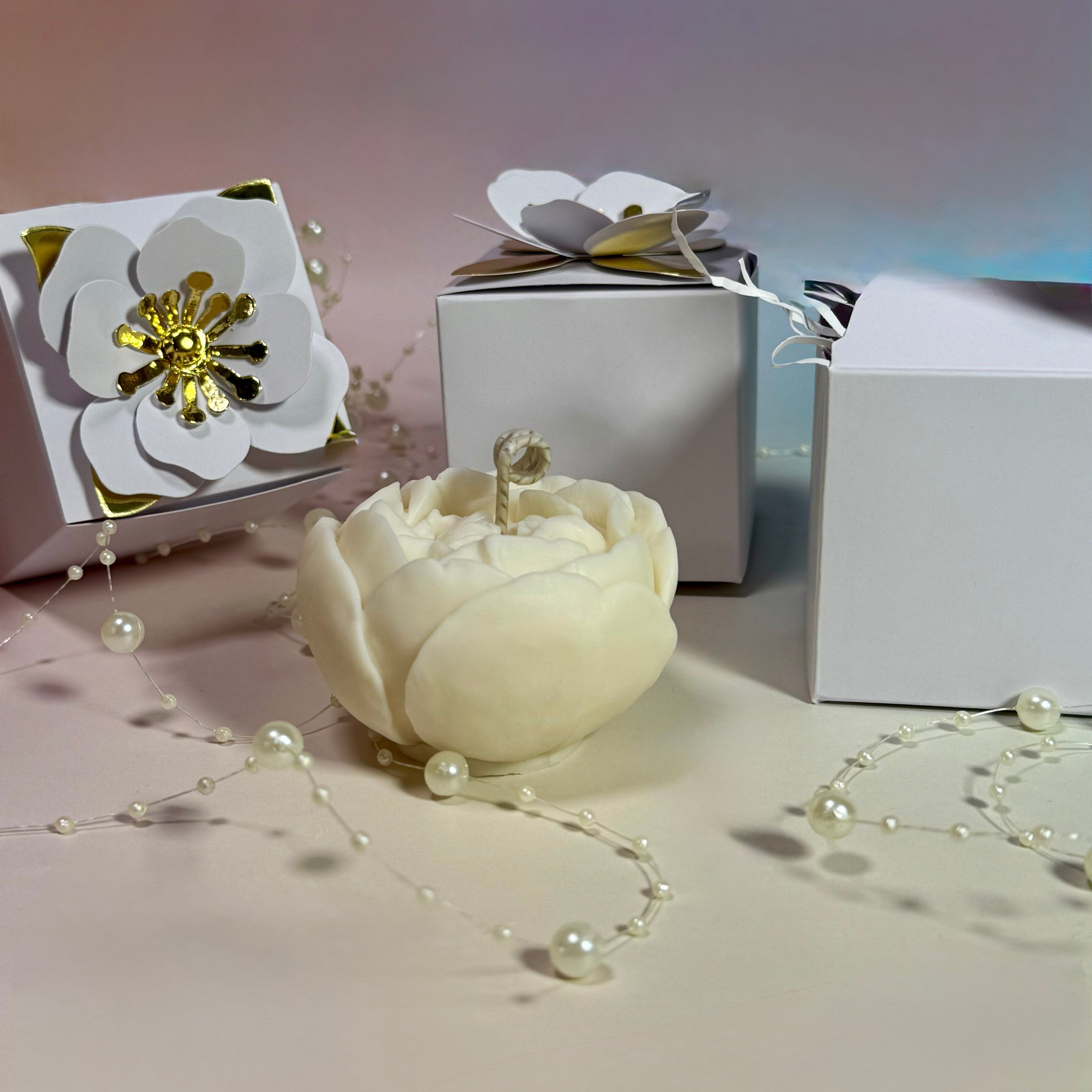 A handcrafted natural white soy wax peony flower candle sitting in front of luxury white gift boxes decorated with 3D paper flowers and delicate pearl strands.