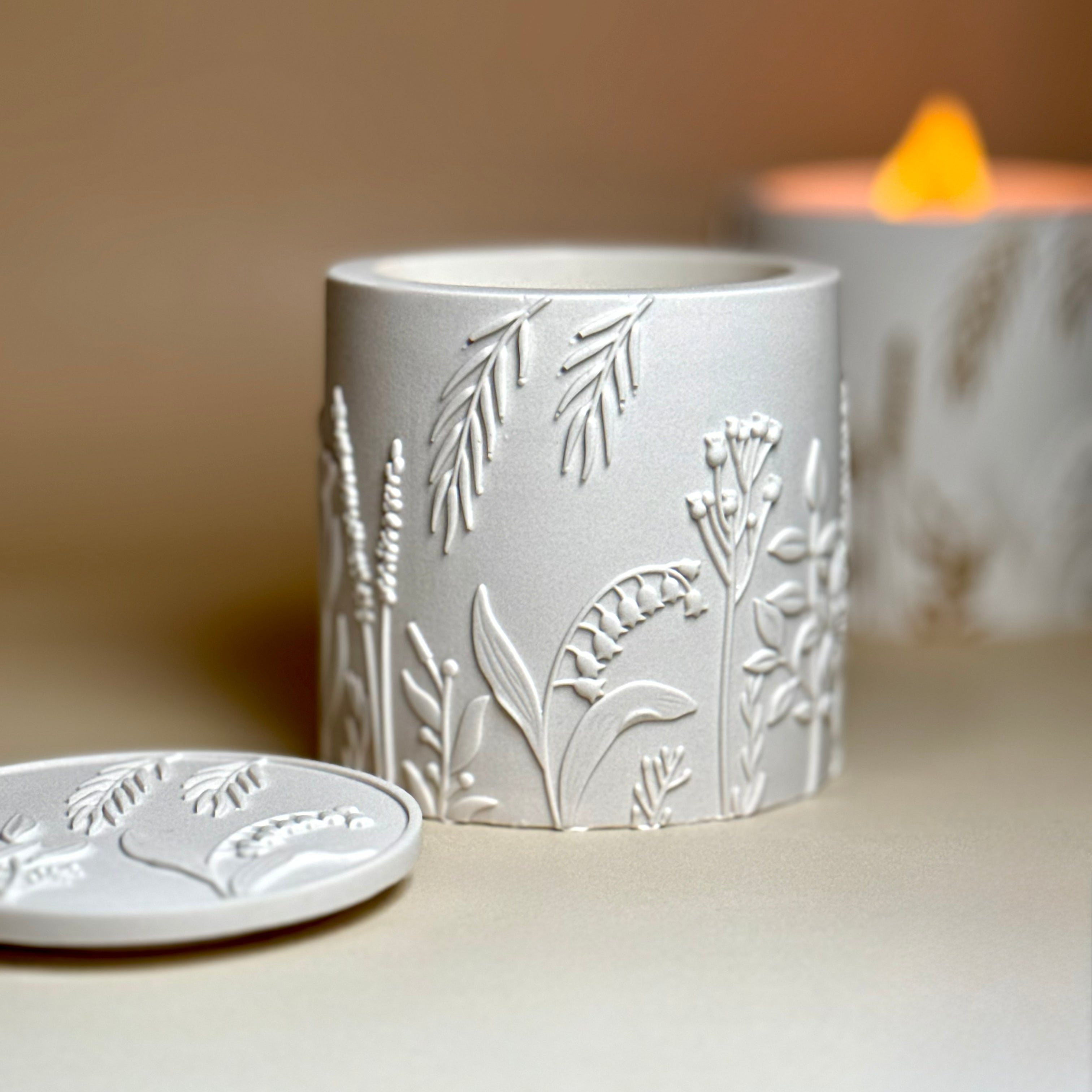 An angle view of an open classic white Jesmonite botanical candle without a lid with its matching detailed botanical relief lid resting beside it.