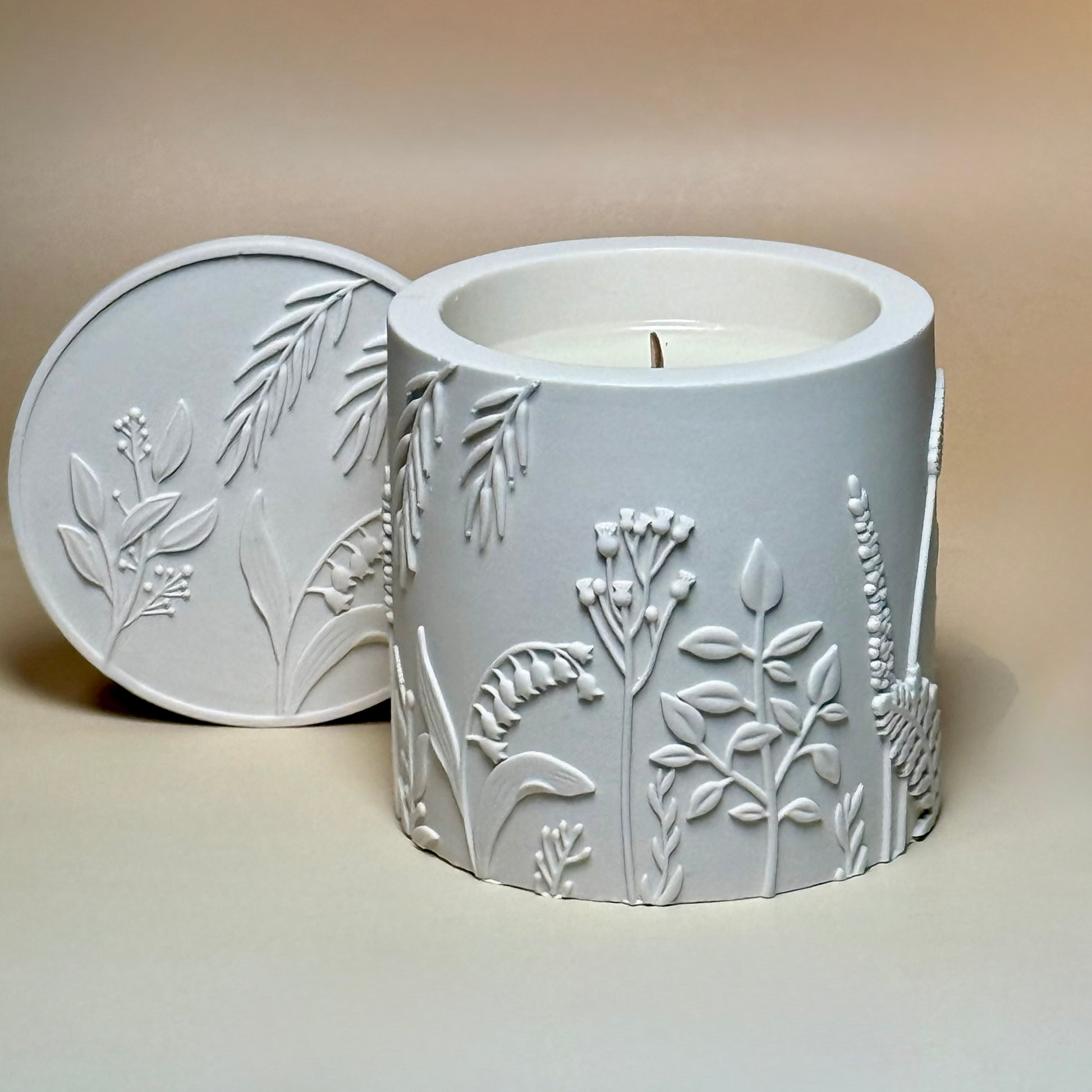 A direct top-down view of an open classic white botanical relief candle, displaying the smooth natural soy wax and wooden wick inside the Jesmonite vessel, with its matching detailed lid resting beside it.