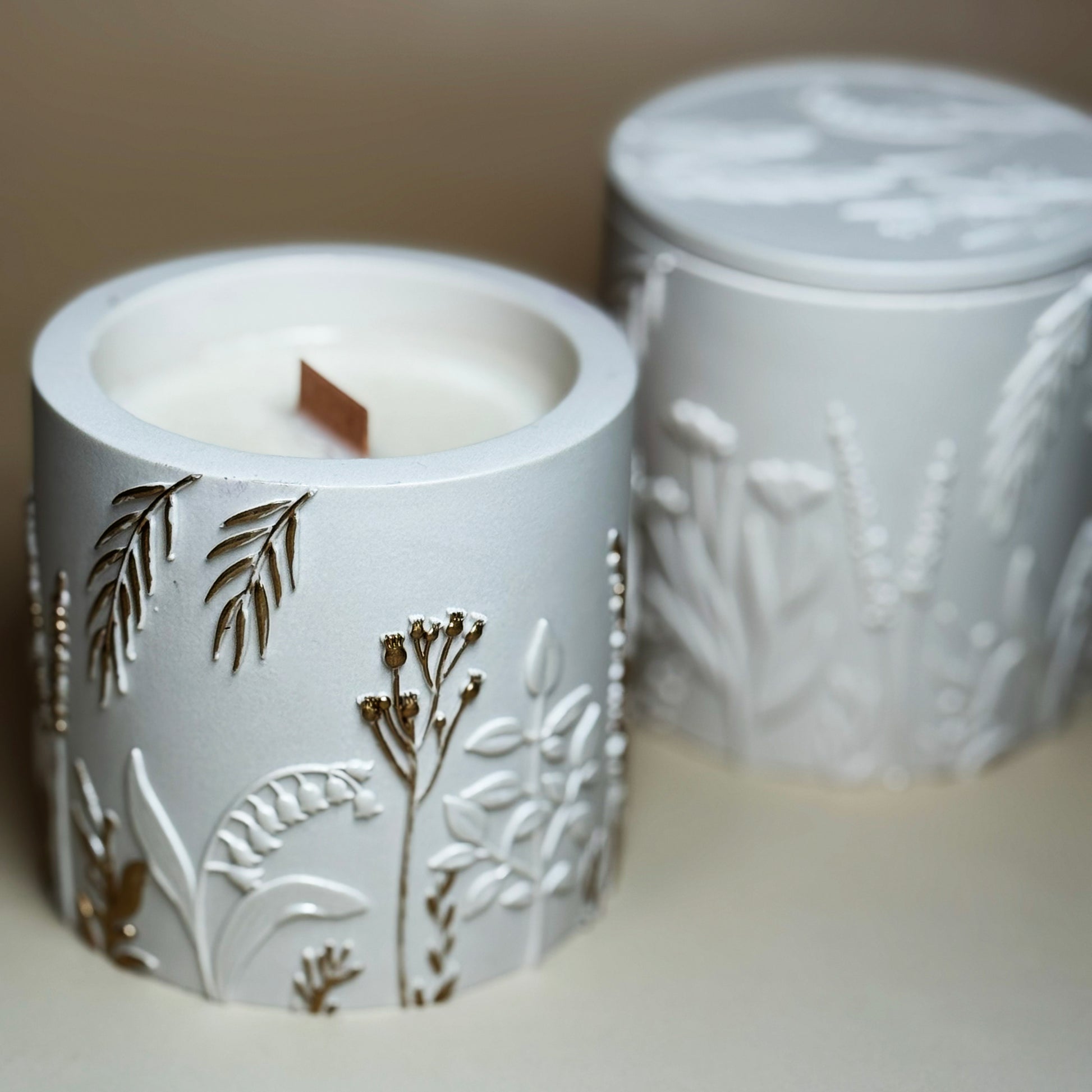 An open hand-painted gold botanical candle with a visible wooden wick, styled in front of a closed classic pearlescent white botanical relief candle jar in soft focus.