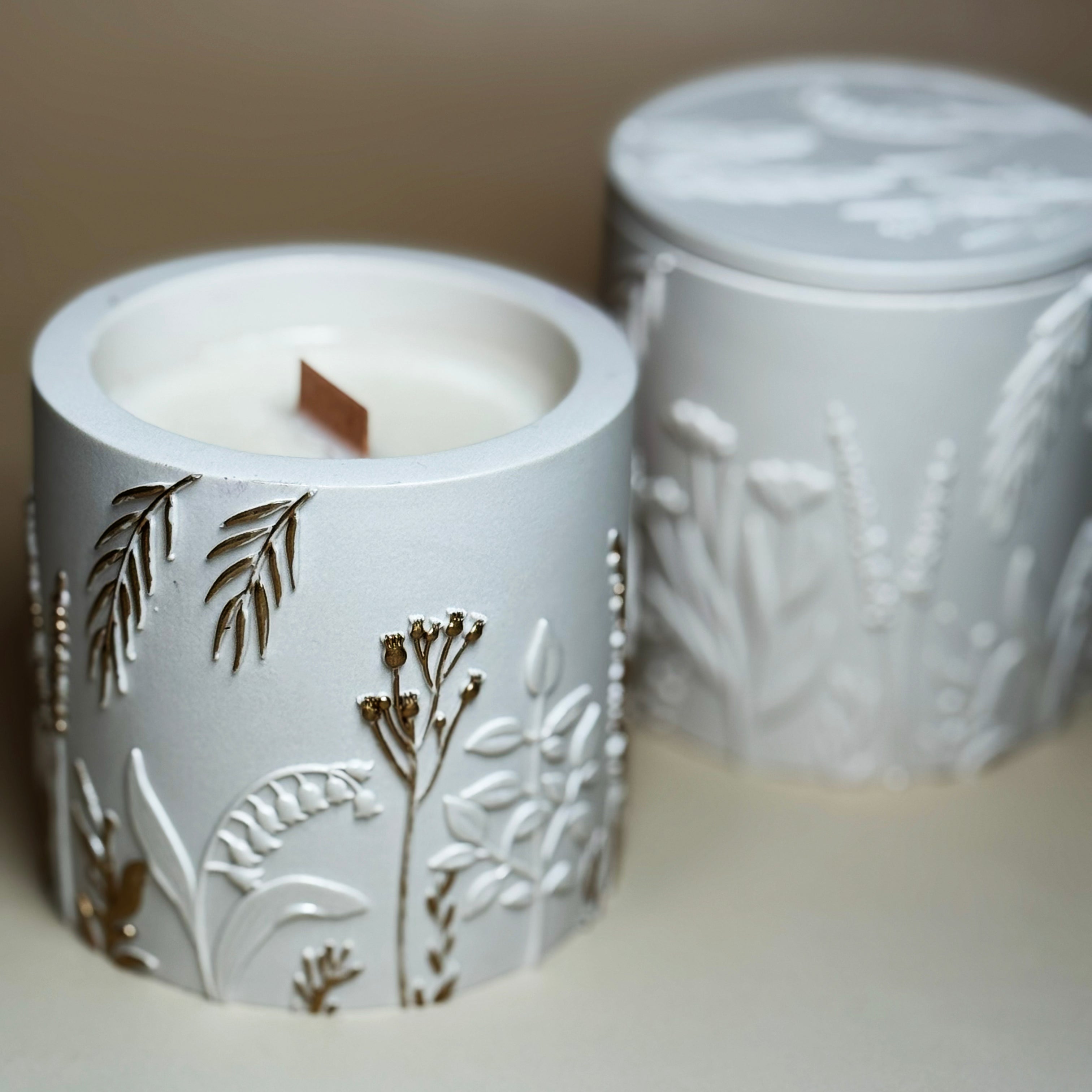 An open hand-painted gold botanical candle with a visible wooden wick, styled in front of a closed classic pearlescent white botanical relief candle jar in soft focus.