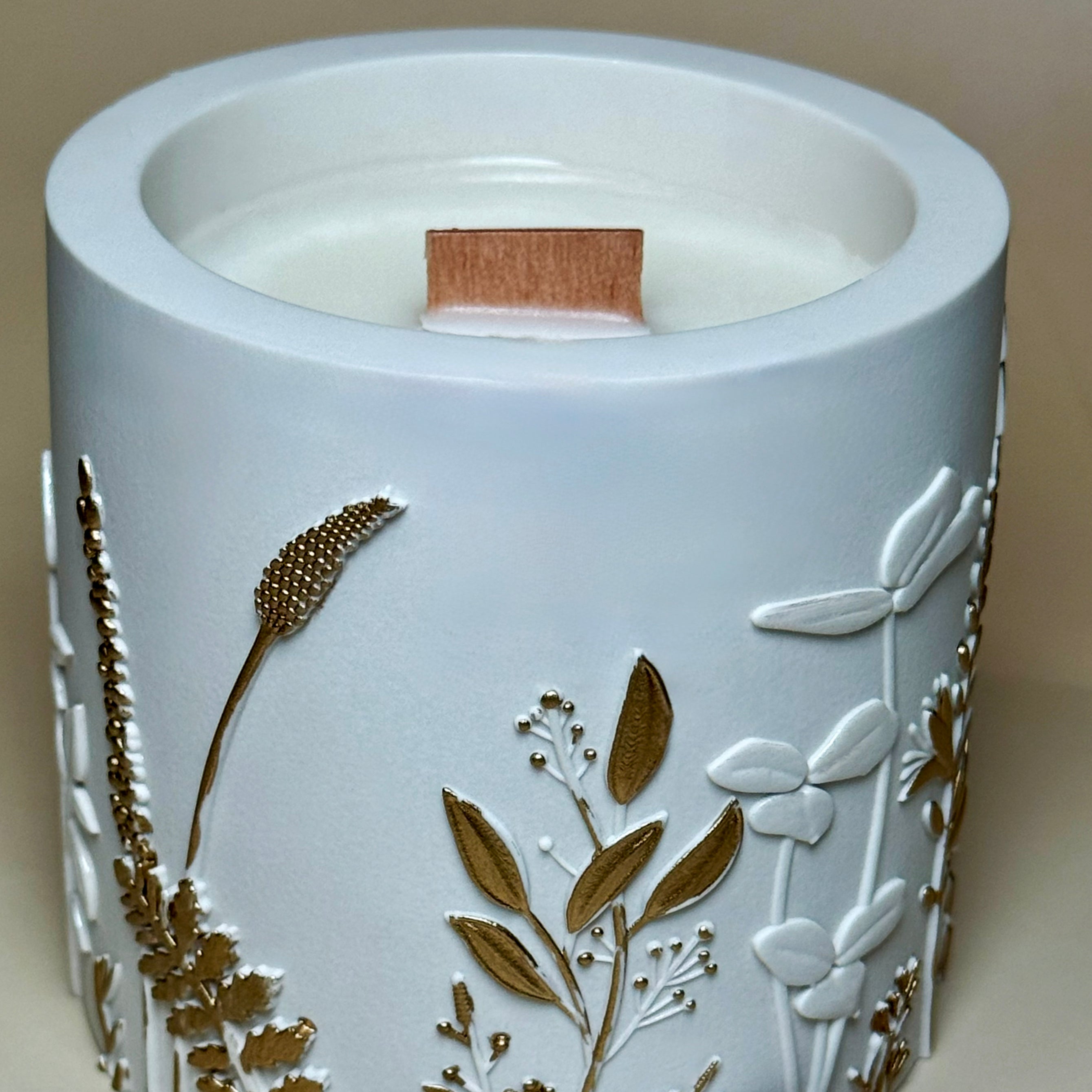 An open hand-painted gold botanical candle showing the smooth soy wax inside, with its detailed floral relief lid resting flat beside the Jesmonite pot.
