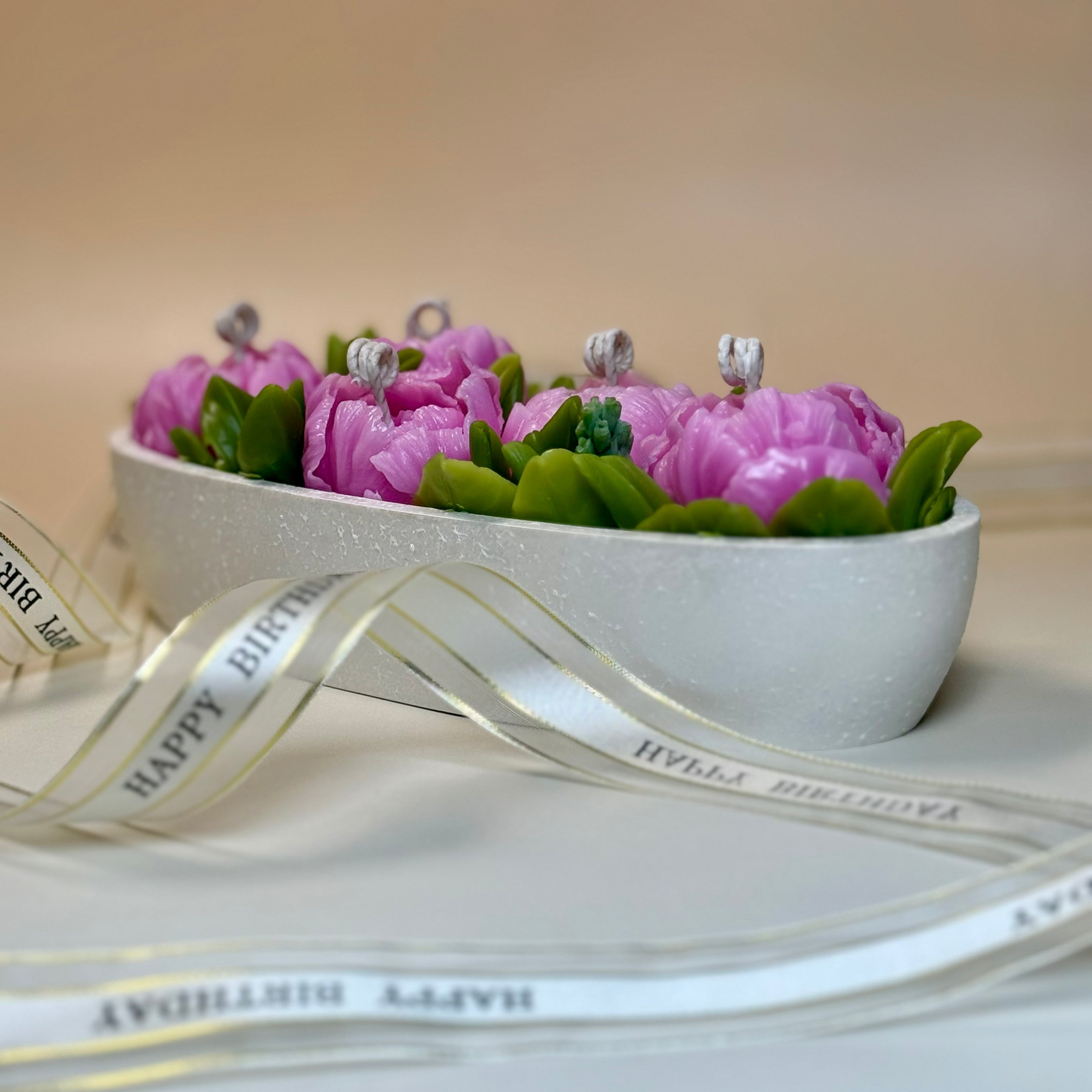Side angle of an elegant oval centrepiece floral candle in a pearl-white stone-textured vessel, surrounded by a luxury gold-edged Happy Birthday ribbon with gift box.