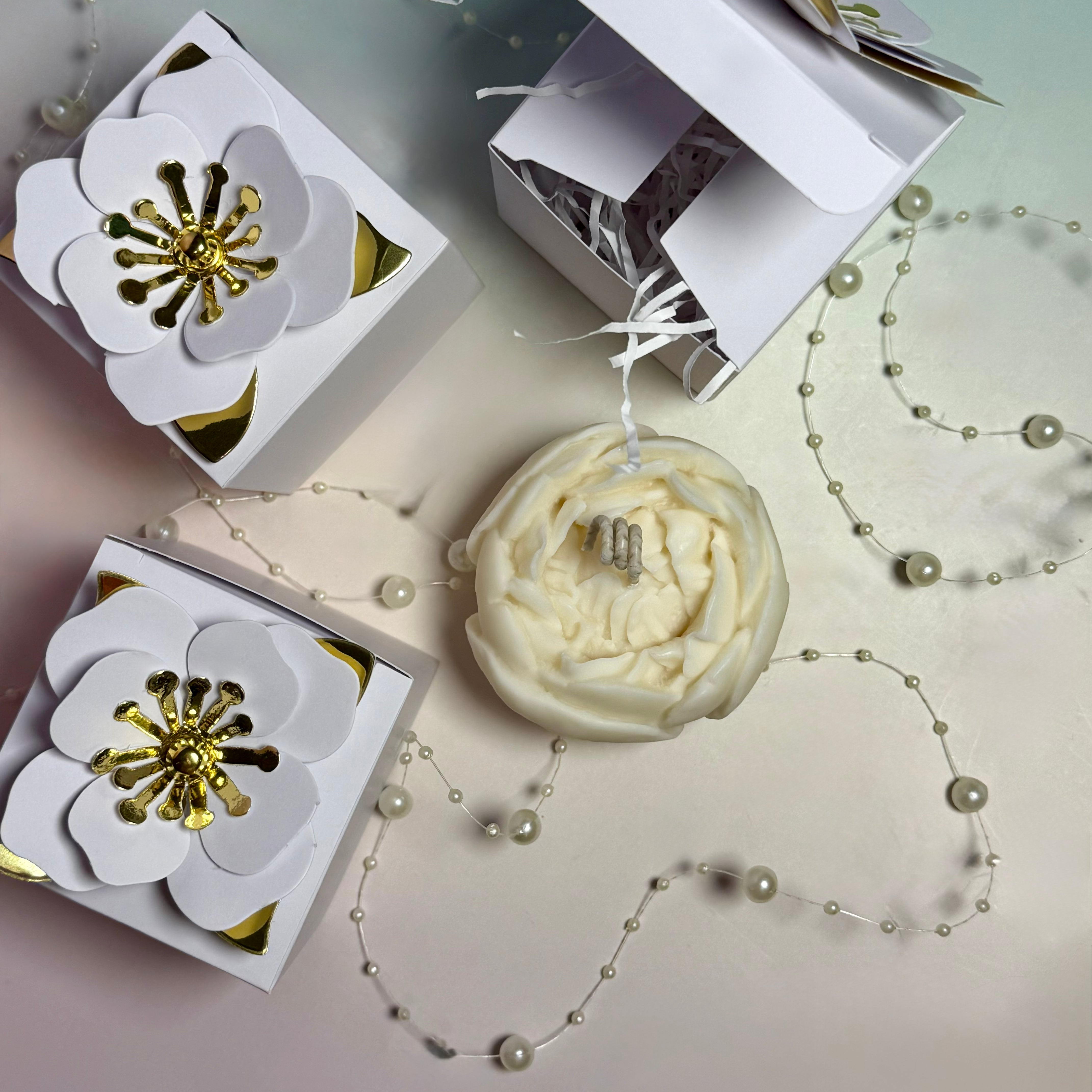 An overhead shot of a detailed cream-coloured peony wax candle surrounded by luxury white gift boxes topped with large 3D paper blooms with gold foil stamens.