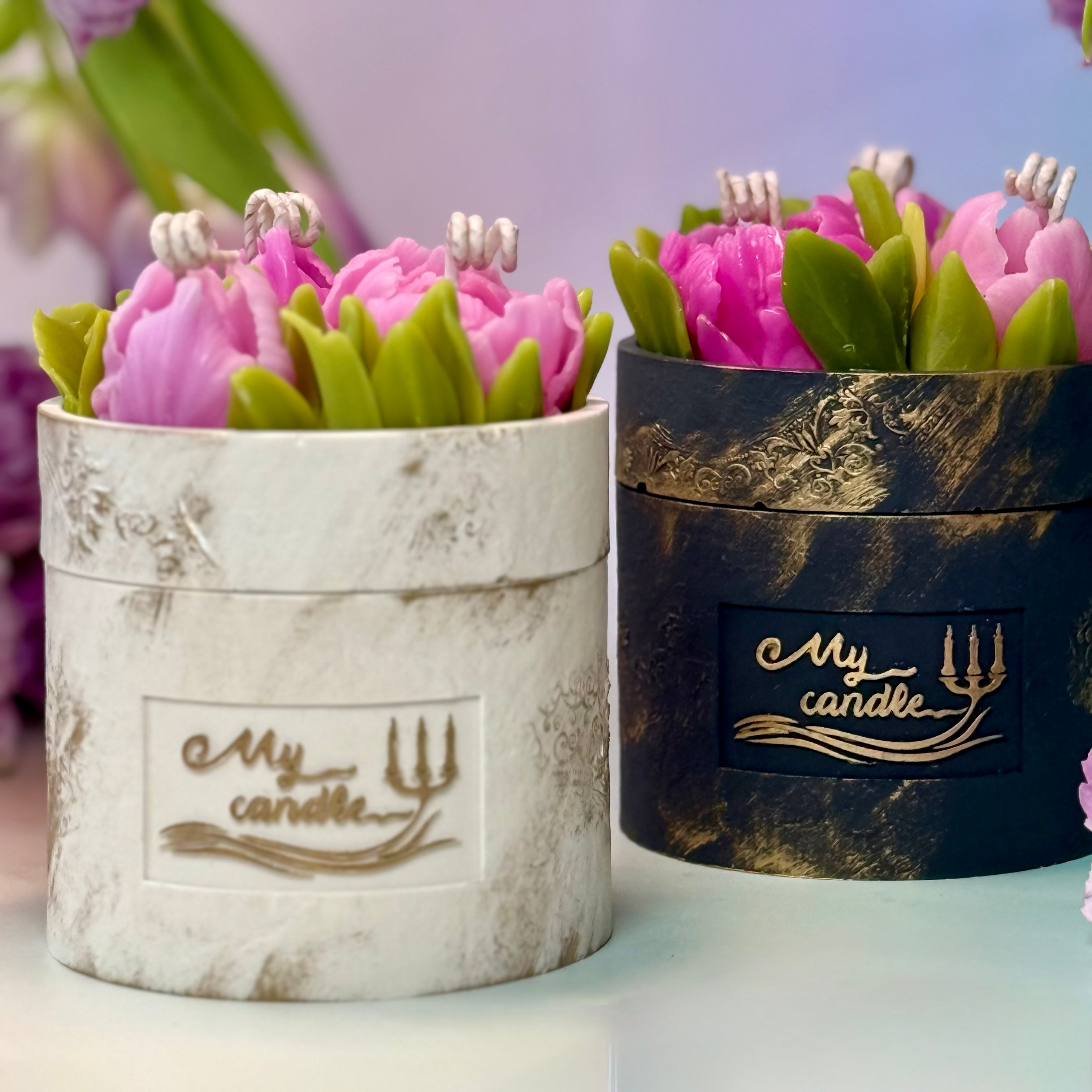 Two artisan flower candle vessels with embossed logos in pearlescent white and black gold patina, topped with hand-moulded purple beeswax tulip bouquets.