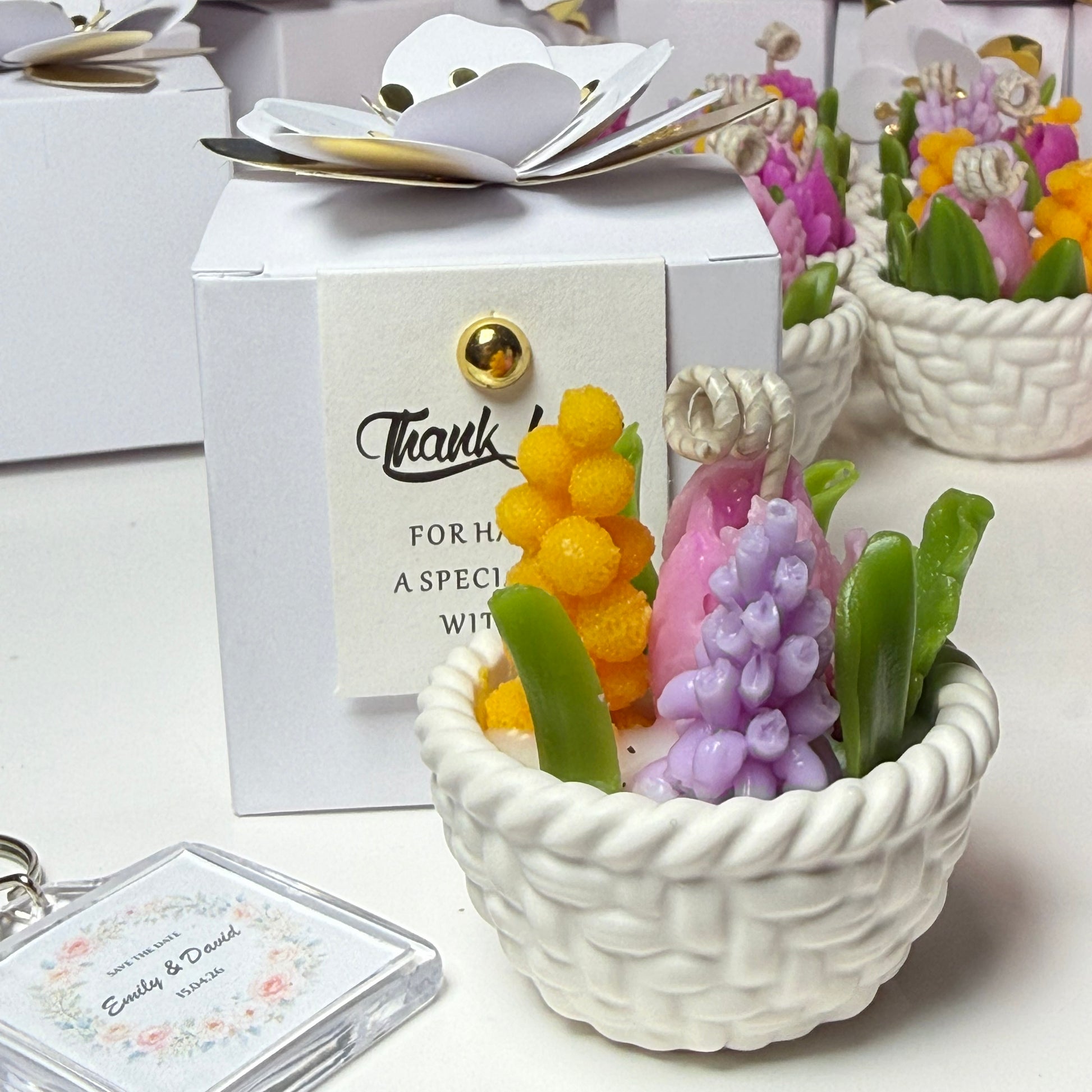 A handcrafted white Jesmonite basket candle filled with colourful purple, pink, and yellow beeswax flowers, presented next to a white gift box with a thank you tag and a personalised wedding keychain.