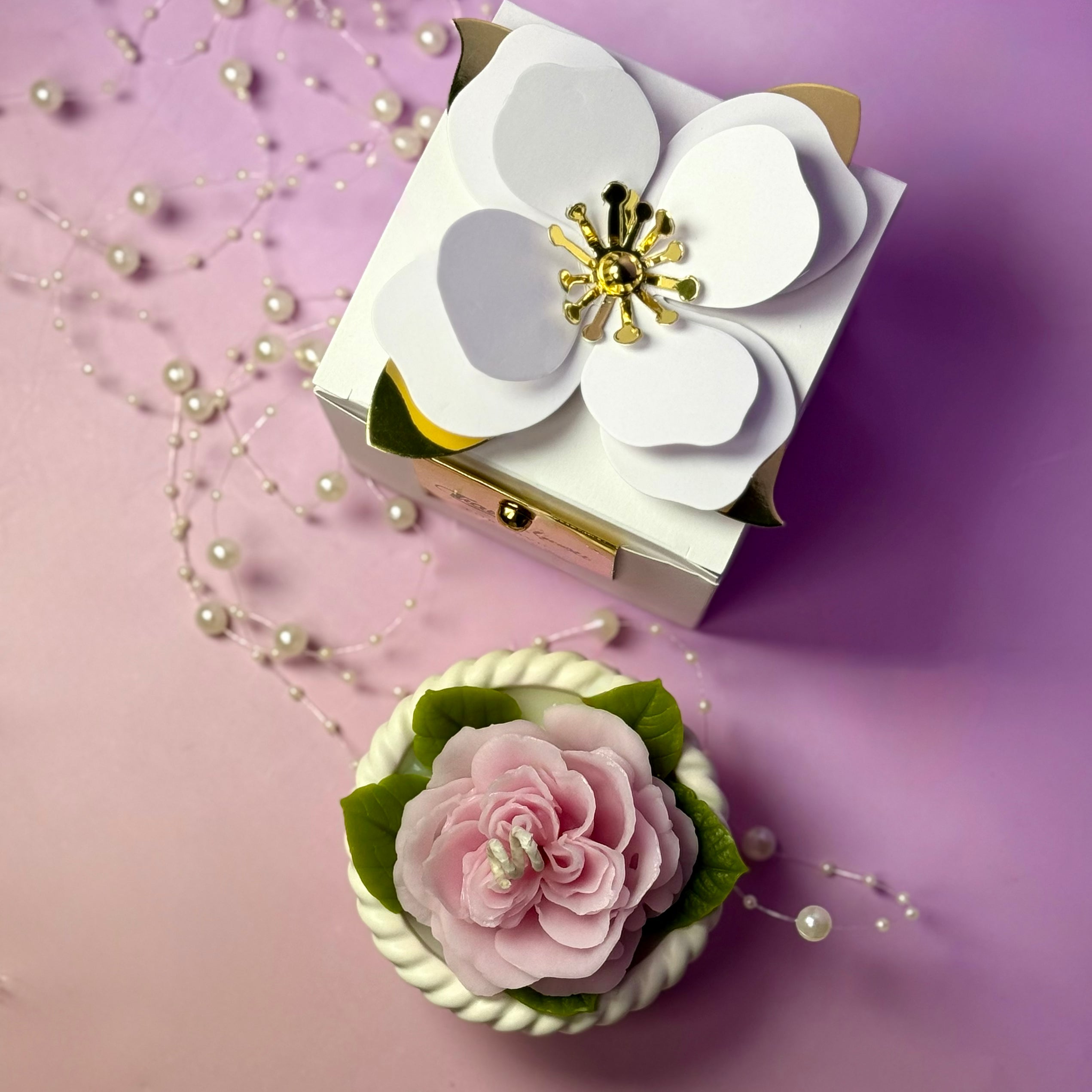 An overhead shot of the closed white gift box featuring a large, artistic 3D white paper magnolia flower on the lid next to the pink rose candle.