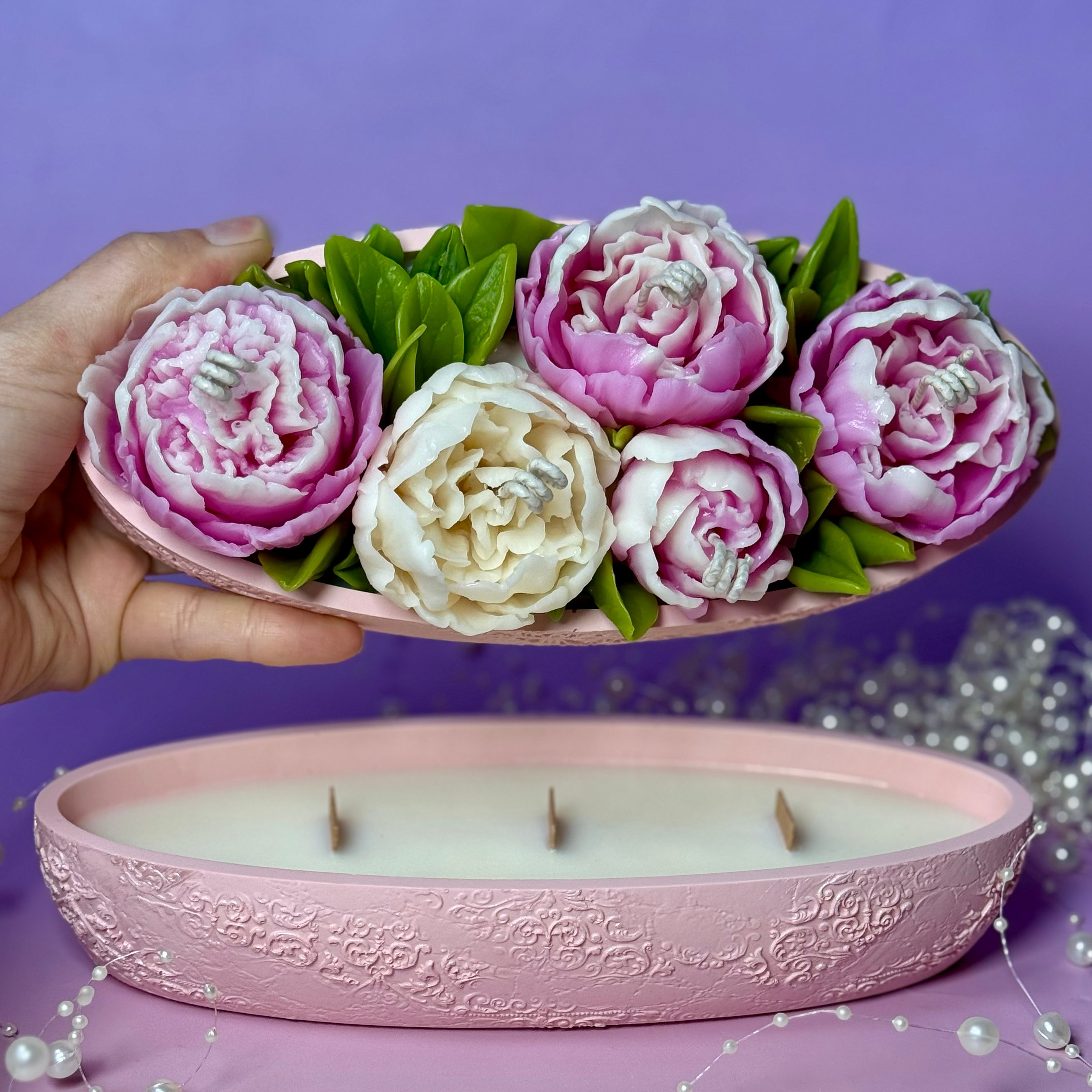 A hand lifting the decorative hand-sculpted beeswax flower lid to reveal the 3-wick soya candle inside the pink textured Jesmonite base pot.