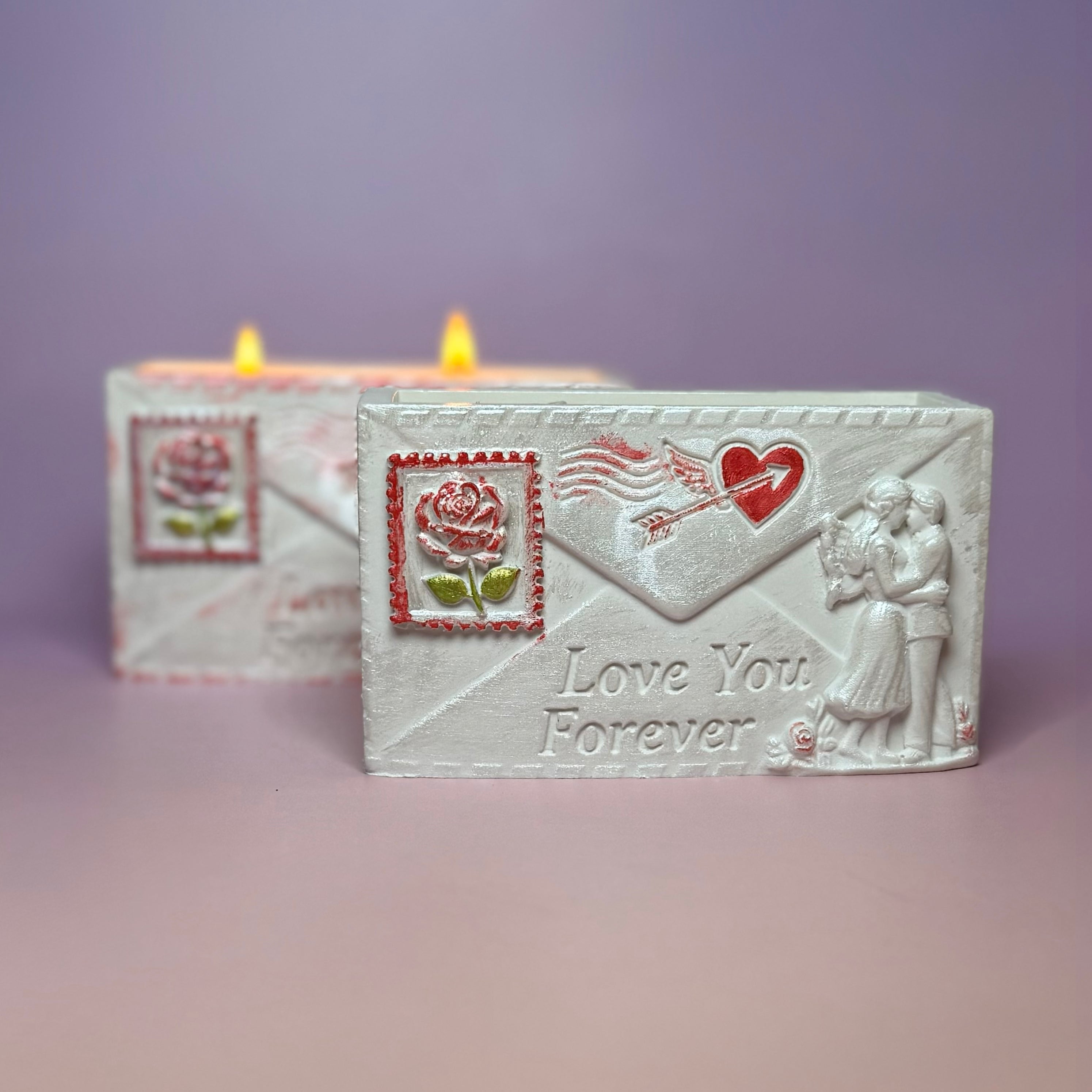 Front view of the unlit pearl-white Love You Forever envelope candle container next to a burning double-wick candle, displaying the embossed winged heart and arrow motif.
