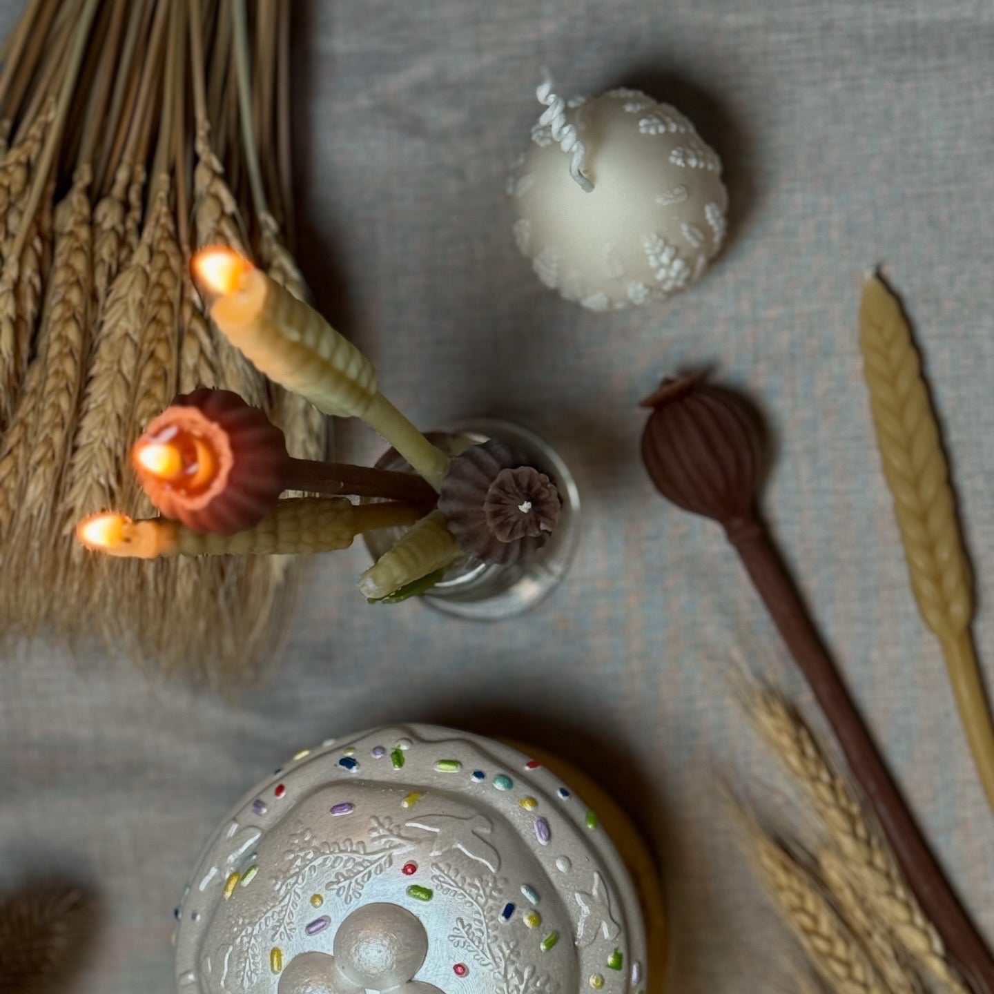 An elegant lifestyle display featuring the Wheat Stalk and Poppy Pod beeswax candles styled as a rustic table centrepiece with dried grass stems and vintage accessories.