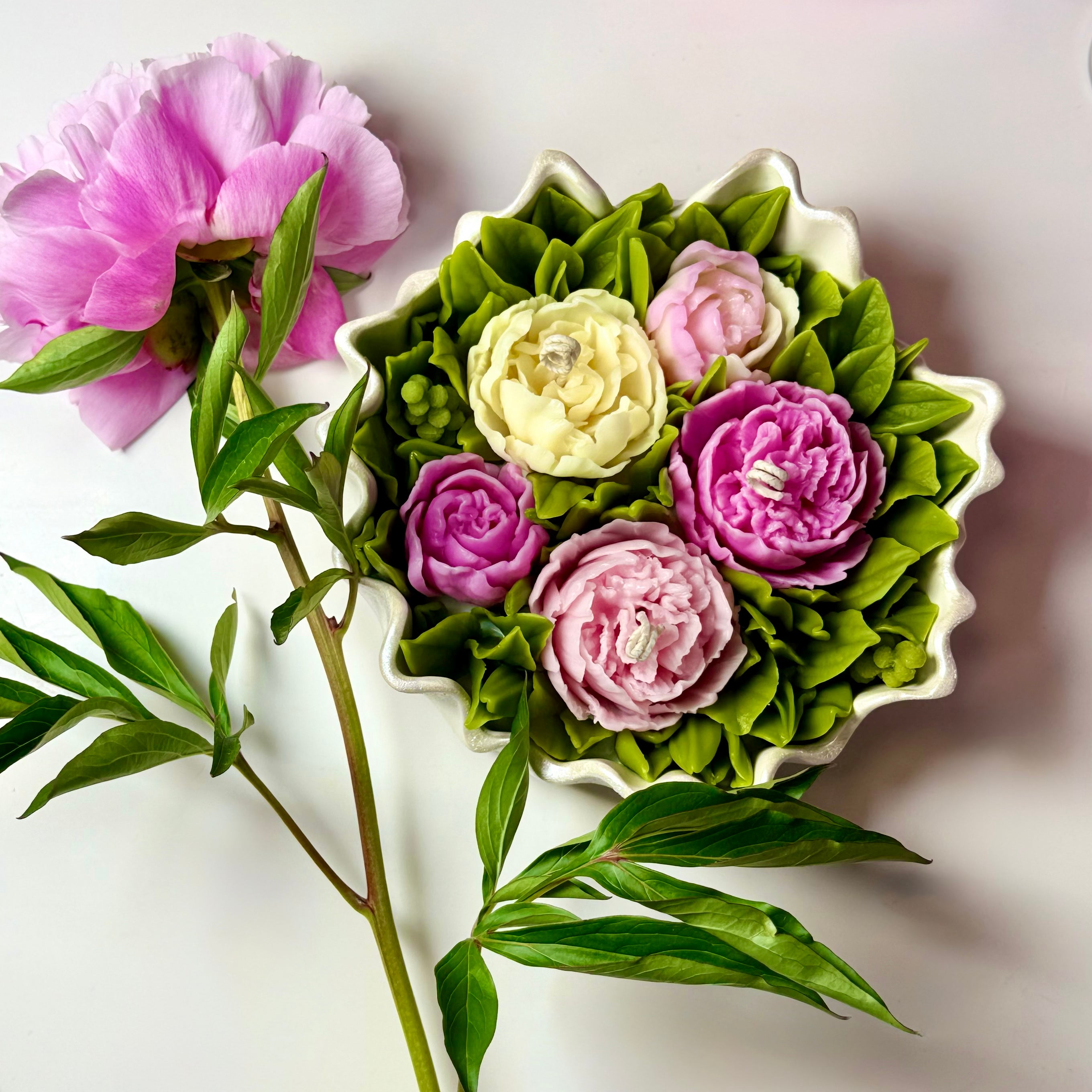 Top-down view of a luxury Sarah Bernhardt peony bouquet candle arrangement next to a real pink peony flower, showcasing realistic hand-poured beeswax petals inside a hand-painted pearl white Jesmonite scalloped bowl.
