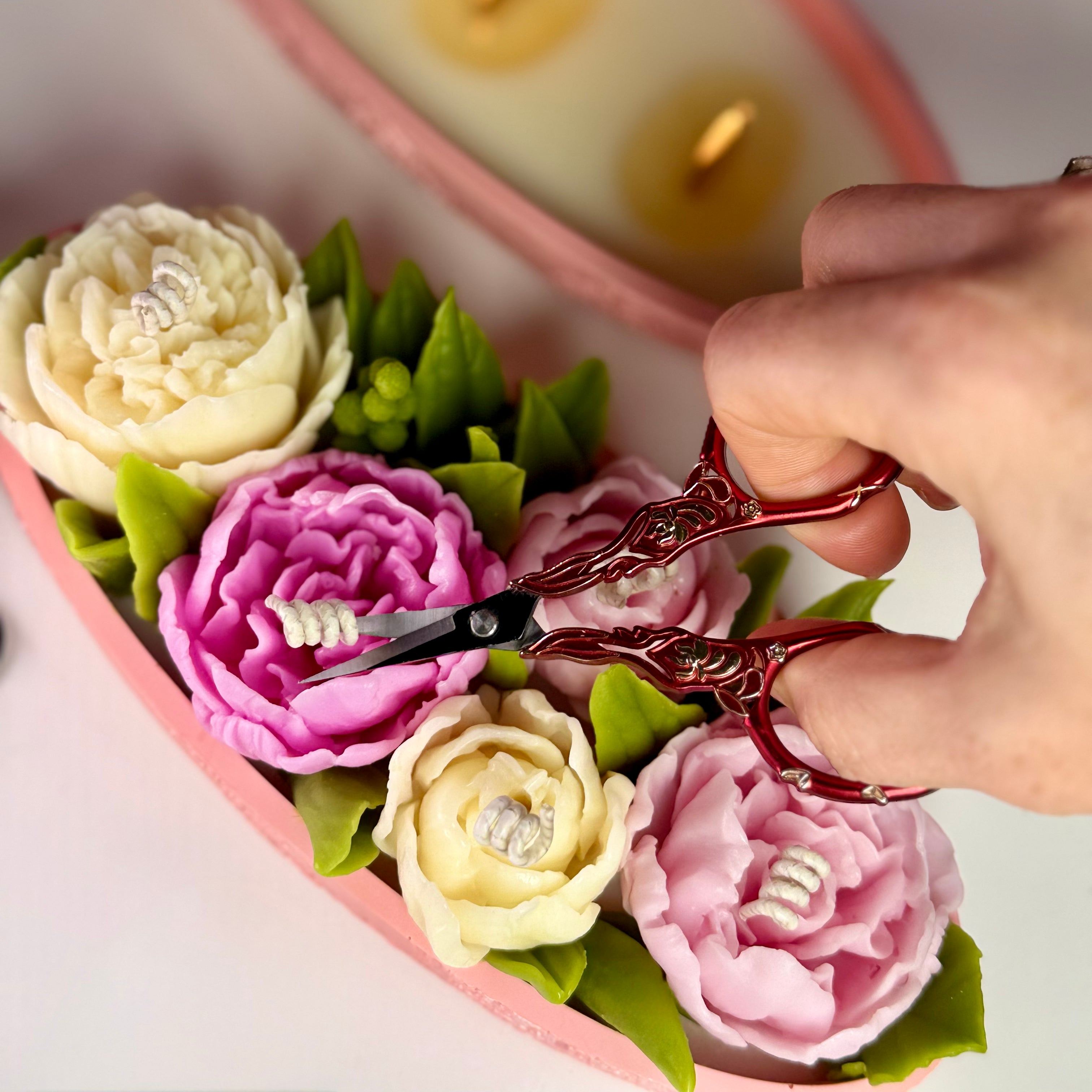 Close-up of ornate red scissors trimming the cotton wicks of highly detailed pink and cream beeswax peony candle flowers.