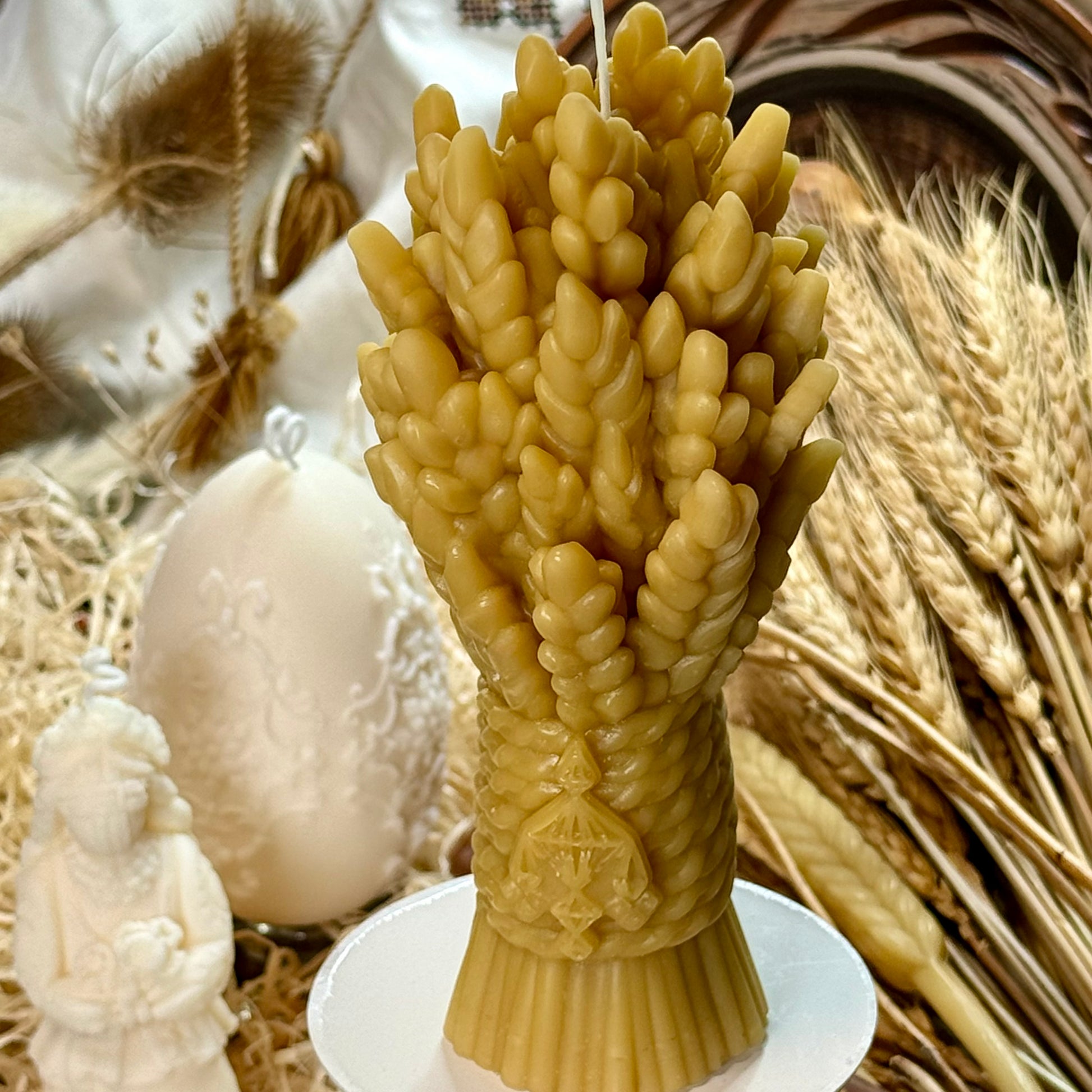 The artisanal Didukh wheat sheaf candle displayed on a white pedestal dish as a table centrepiece, set against a warm rustic backdrop of dried grasses and a white patterned Easter egg.