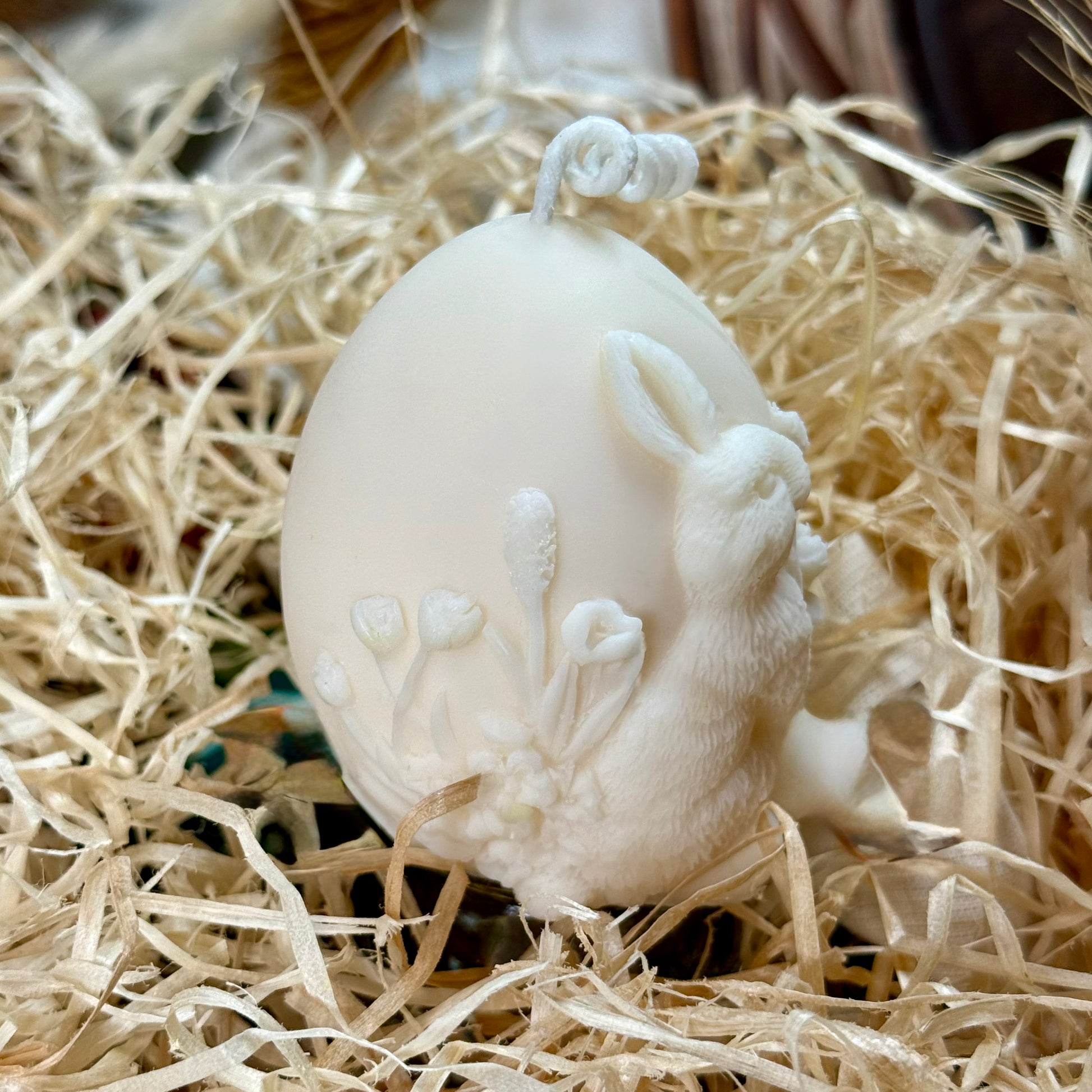 Side view of a hand-poured egg shaped candle featuring a detailed relief of a rabbit and spring wildflowers, sitting in shredded paper packing material as a minimalist Easter decoration.