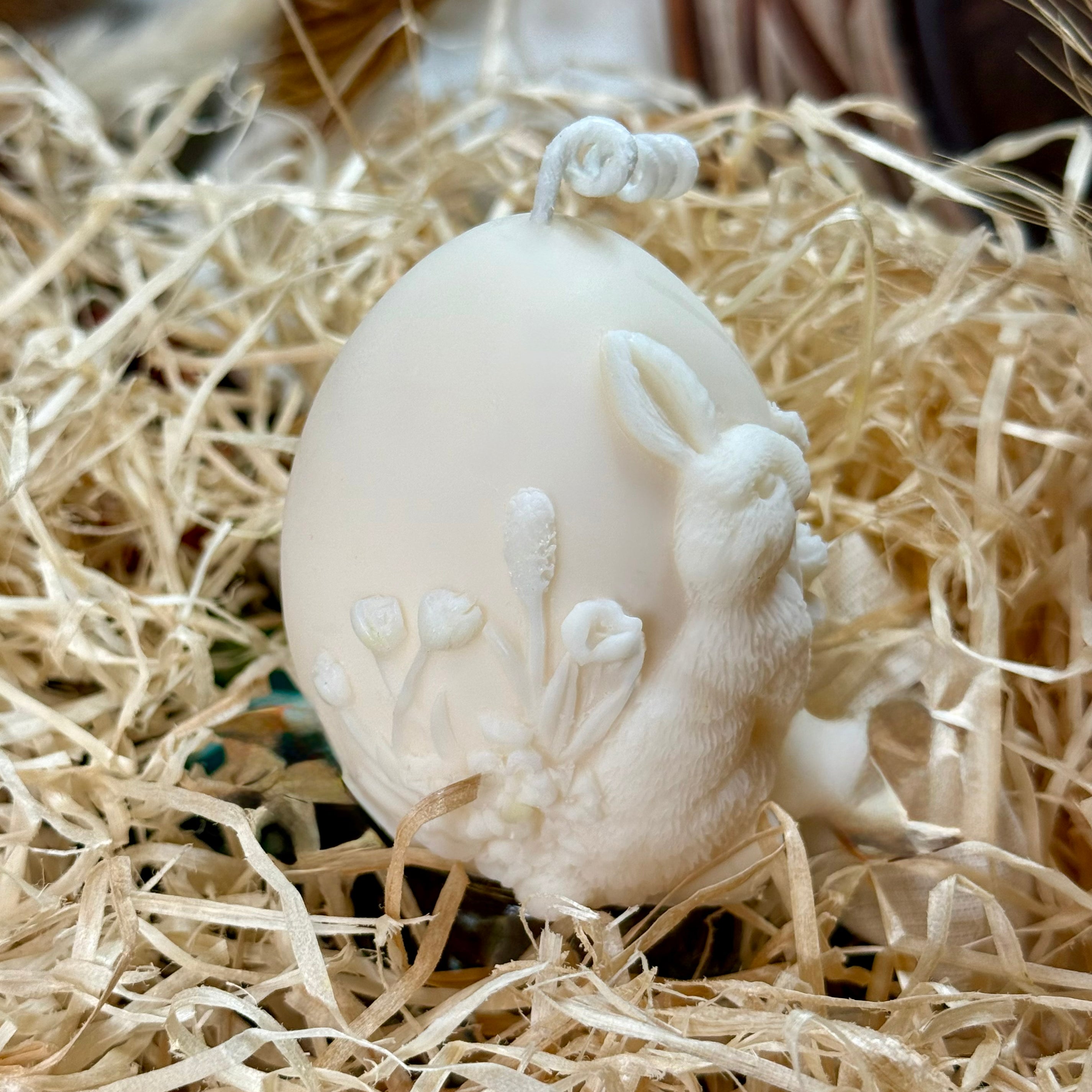 Side view of a hand-poured egg shaped candle featuring a detailed relief of a rabbit and spring wildflowers, sitting in shredded paper packing material as a minimalist Easter decoration.