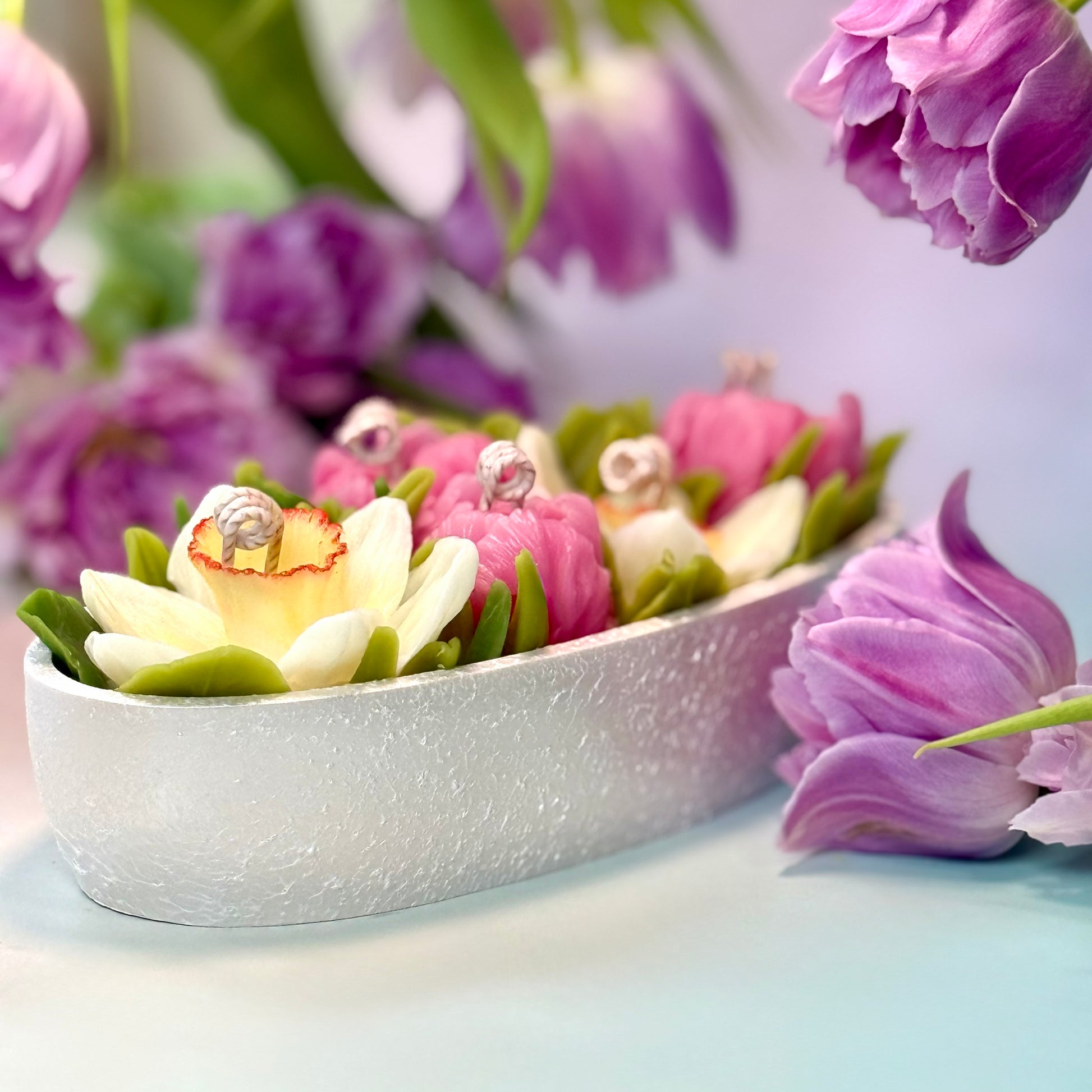 Front macro shot of a white stone-textured oval Jesmonite vessel holding decorative 3D floral candles, surrounded by purple tulip accents with gift box.