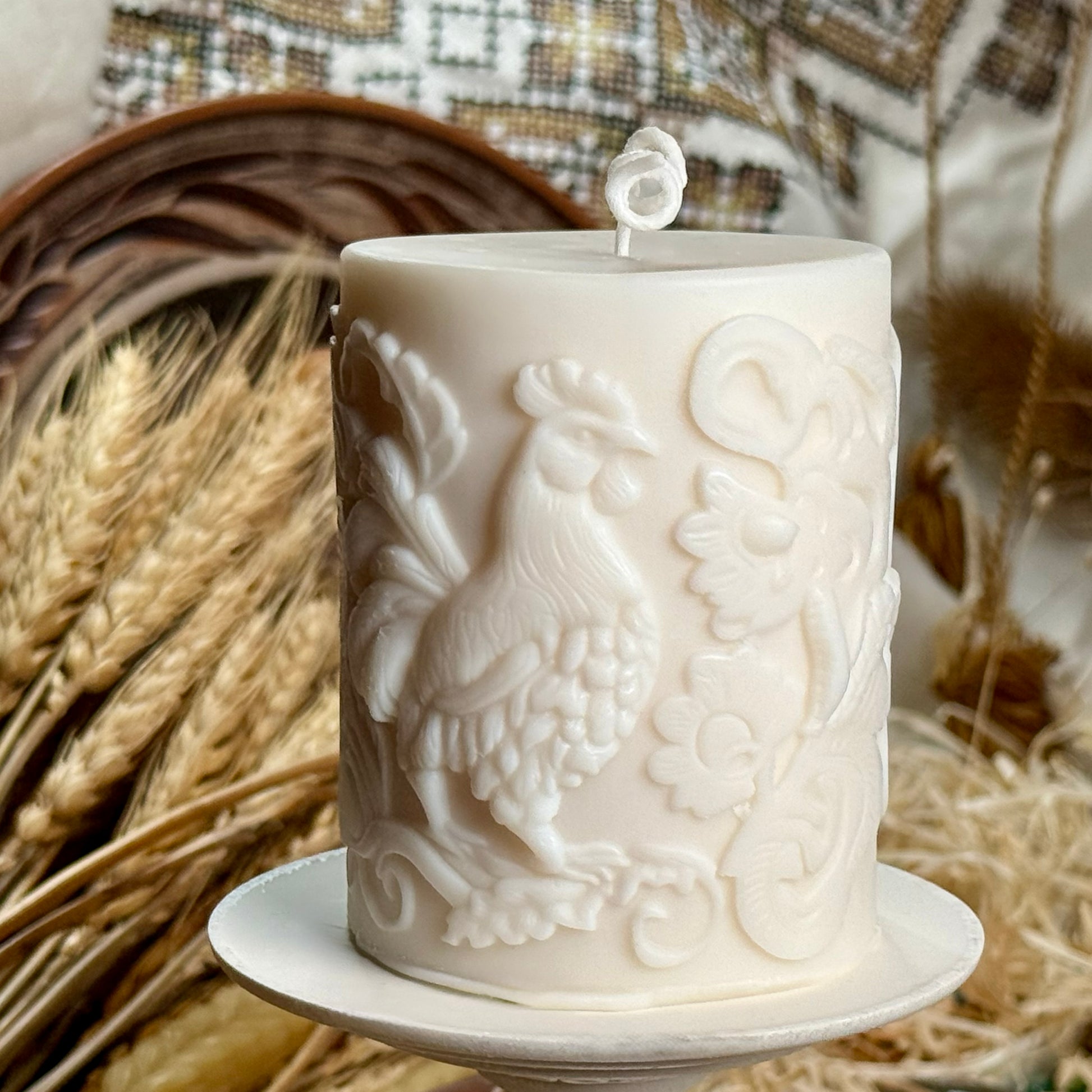 Alt Text: A front-facing view of the folk-art inspired rooster soy wax candle displayed on a white stand, showing the prominent bird motif and elegant curled cotton wick against a rustic country house backdrop.