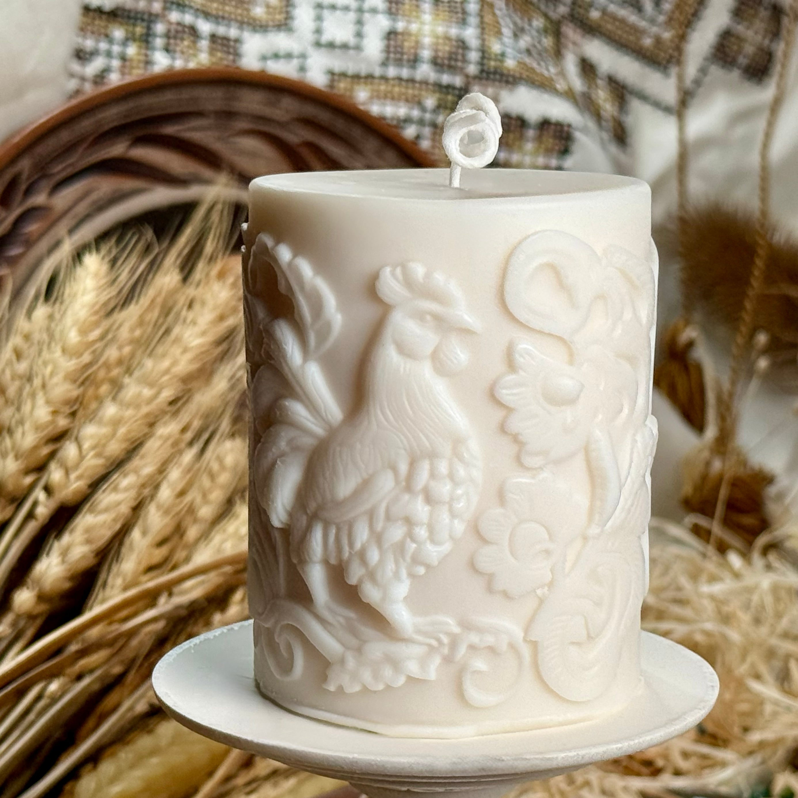 Alt Text: A front-facing view of the folk-art inspired rooster soy wax candle displayed on a white stand, showing the prominent bird motif and elegant curled cotton wick against a rustic country house backdrop.