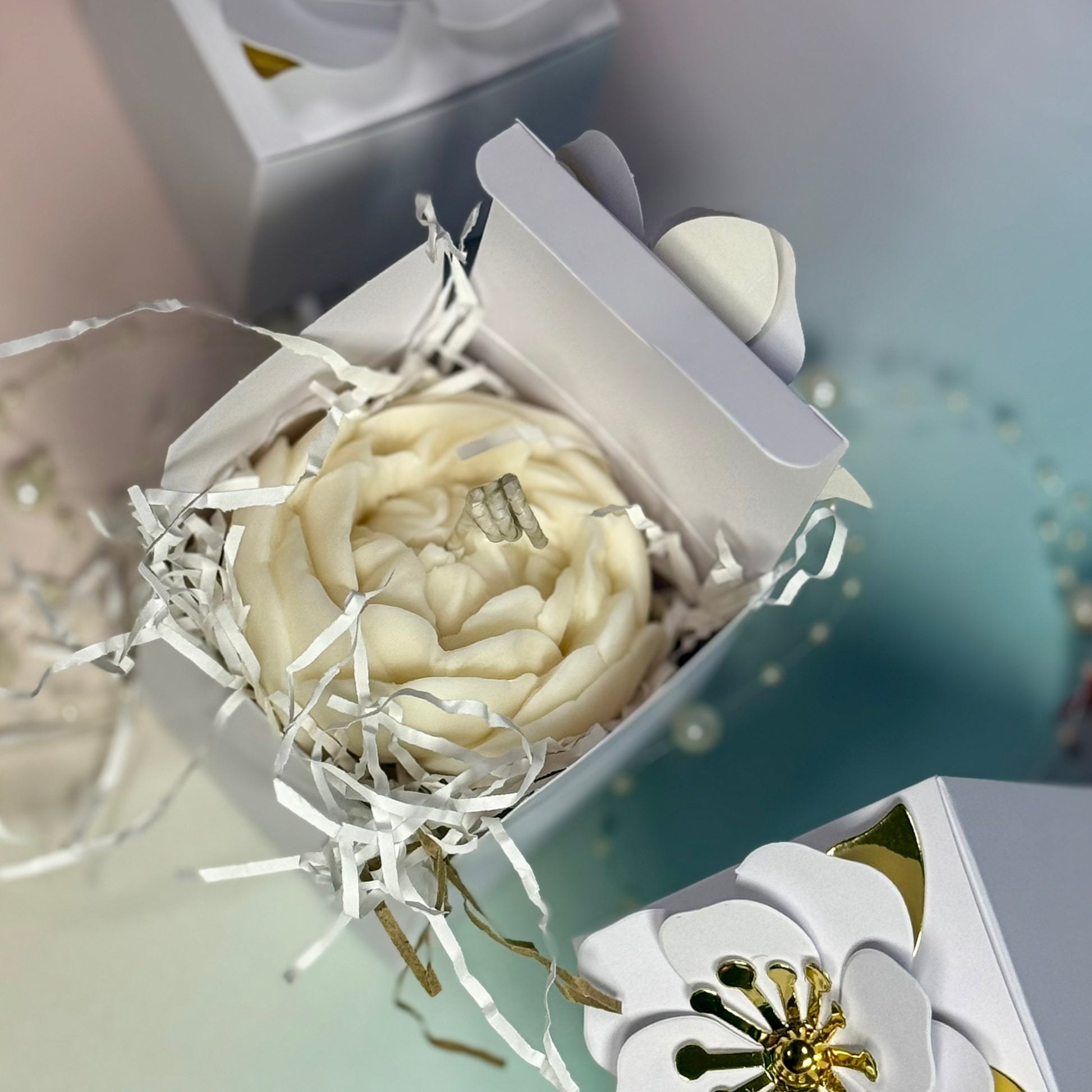 A top-down perspective of an open luxury wedding favour packaging box, highlighting the intricate, clean botanical lines of a hand-poured white peony candle topper.