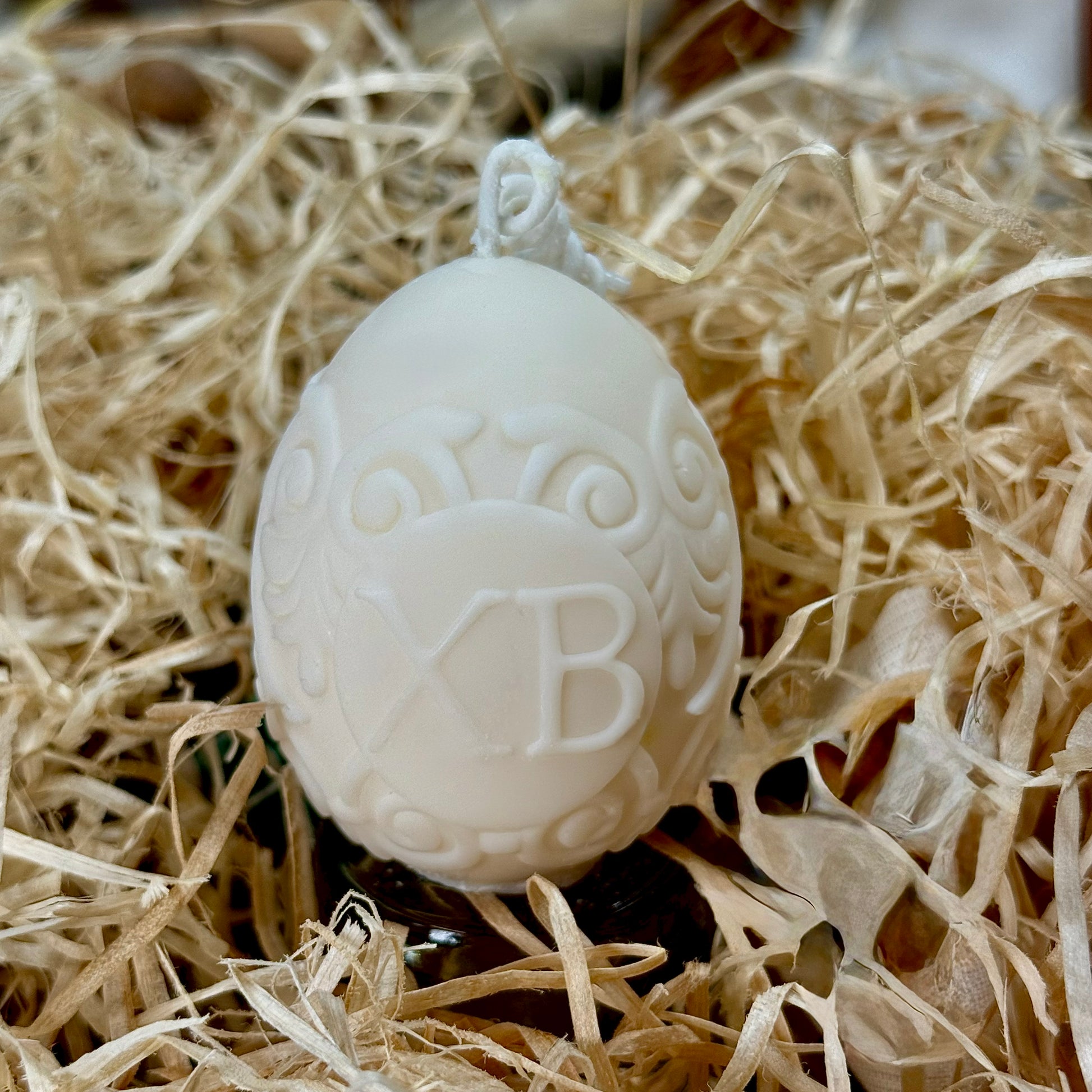 An intricate natural ivory soy wax Easter egg candle featuring the traditional Slavic greeting XB and swirling relief ornaments, resting in a bed of rustic wood shred packaging.