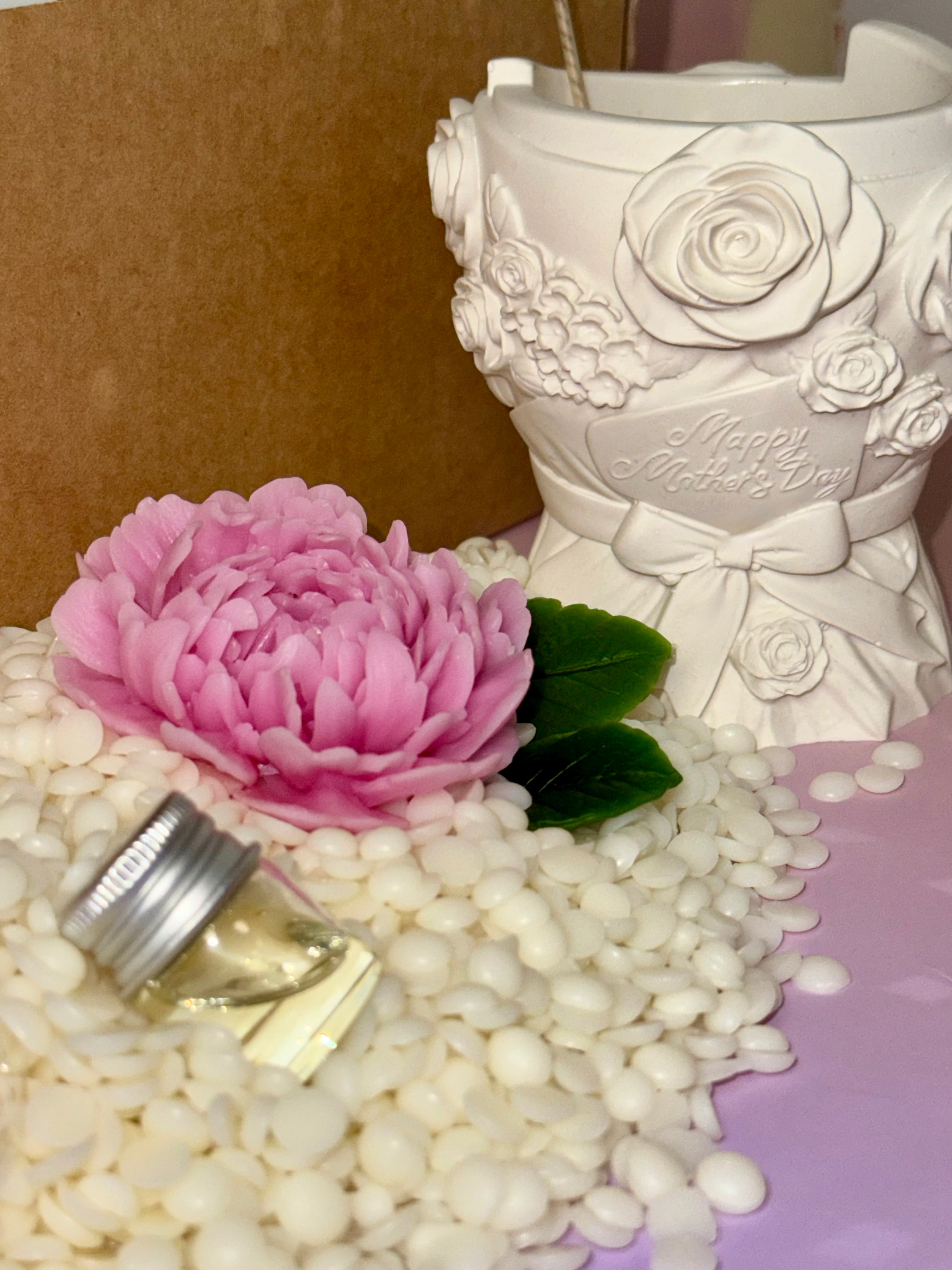 A macro perspective focusing on the unpainted white bouquet vessel with a sculpted Happy Mother's Day inscription, sitting next to a glass bottle of scent oil and a pink beeswax peony flower.