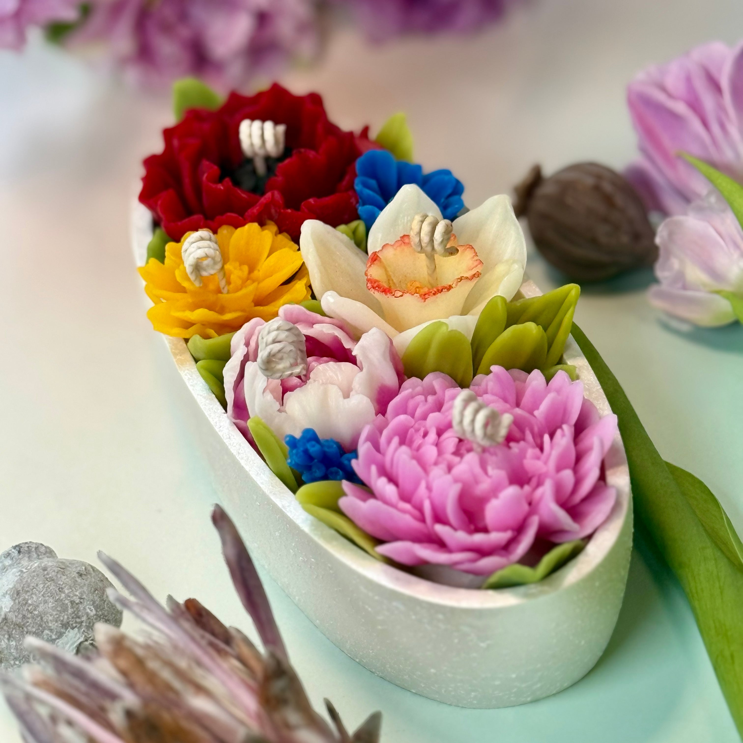 High-angle close-up of an oval floral candle showcasing a stunning mix of a deep red poppy, bright yellow daffodil, and pink beeswax peony blooms with gift box.