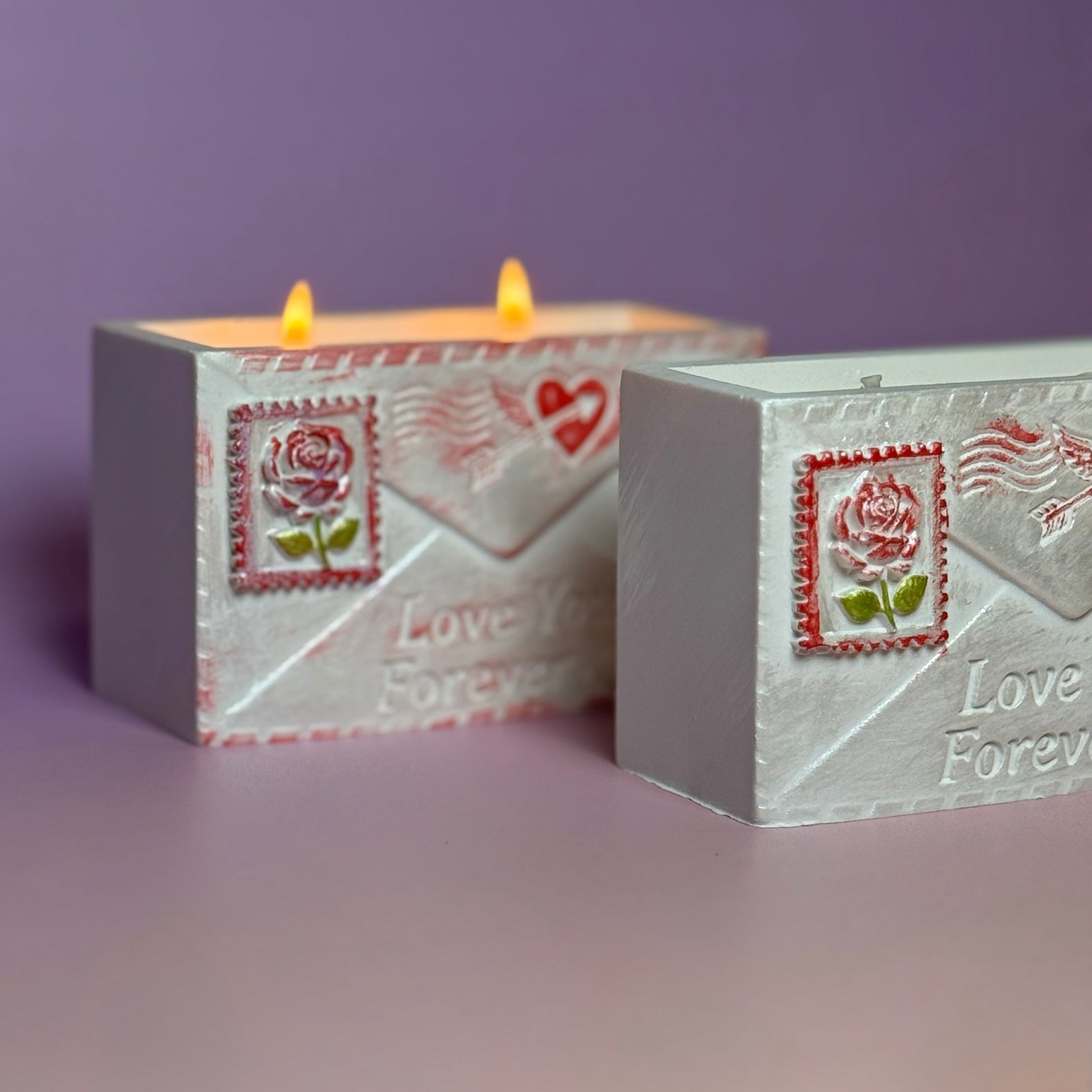 Side angle view of two Jesmonite love letter candle vessels on a purple background, highlighting the clean rectangular lines and the hand-painted rose postage stamp detailing.