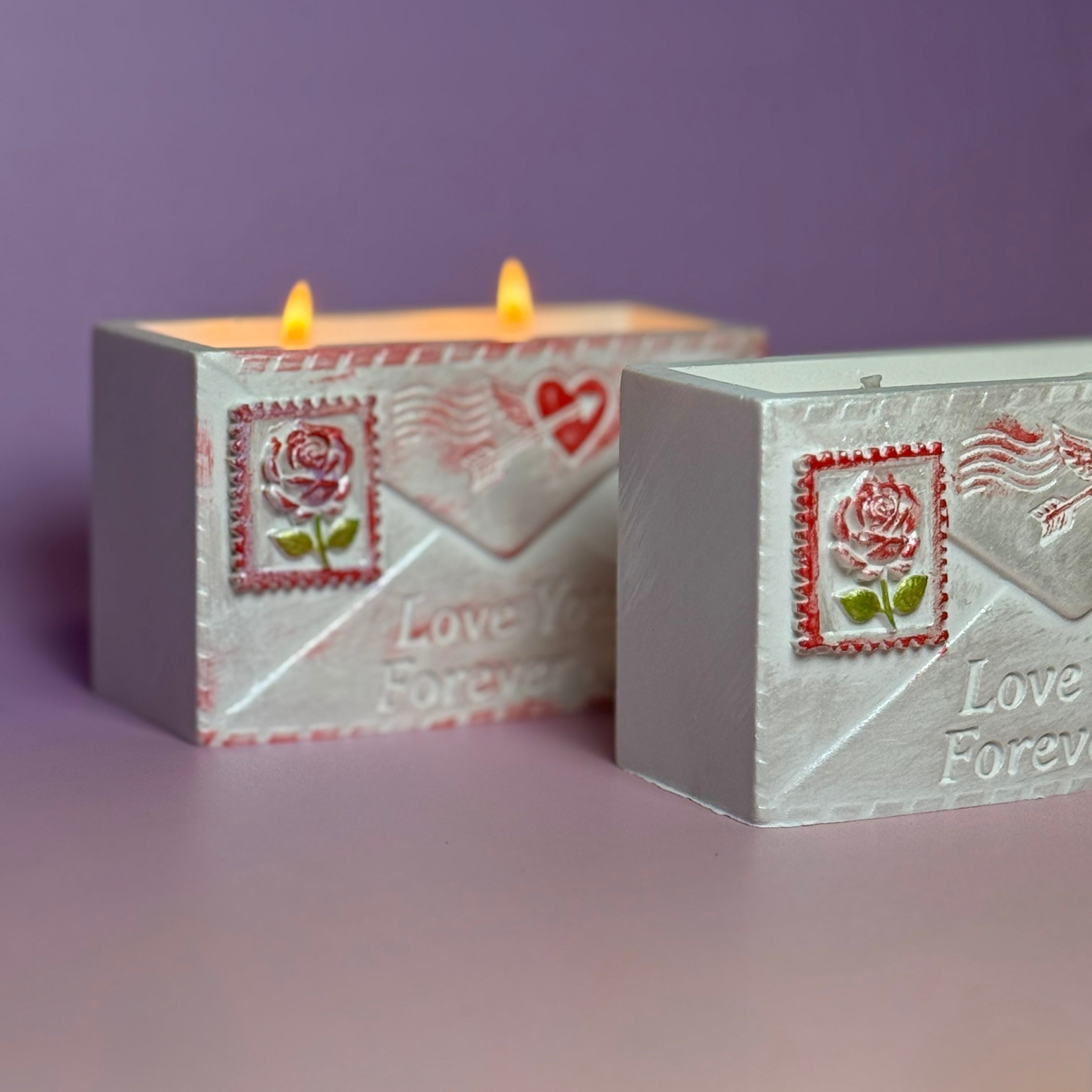 Side angle view of two Jesmonite love letter candle vessels on a purple background, highlighting the clean rectangular lines and the hand-painted rose postage stamp detailing.