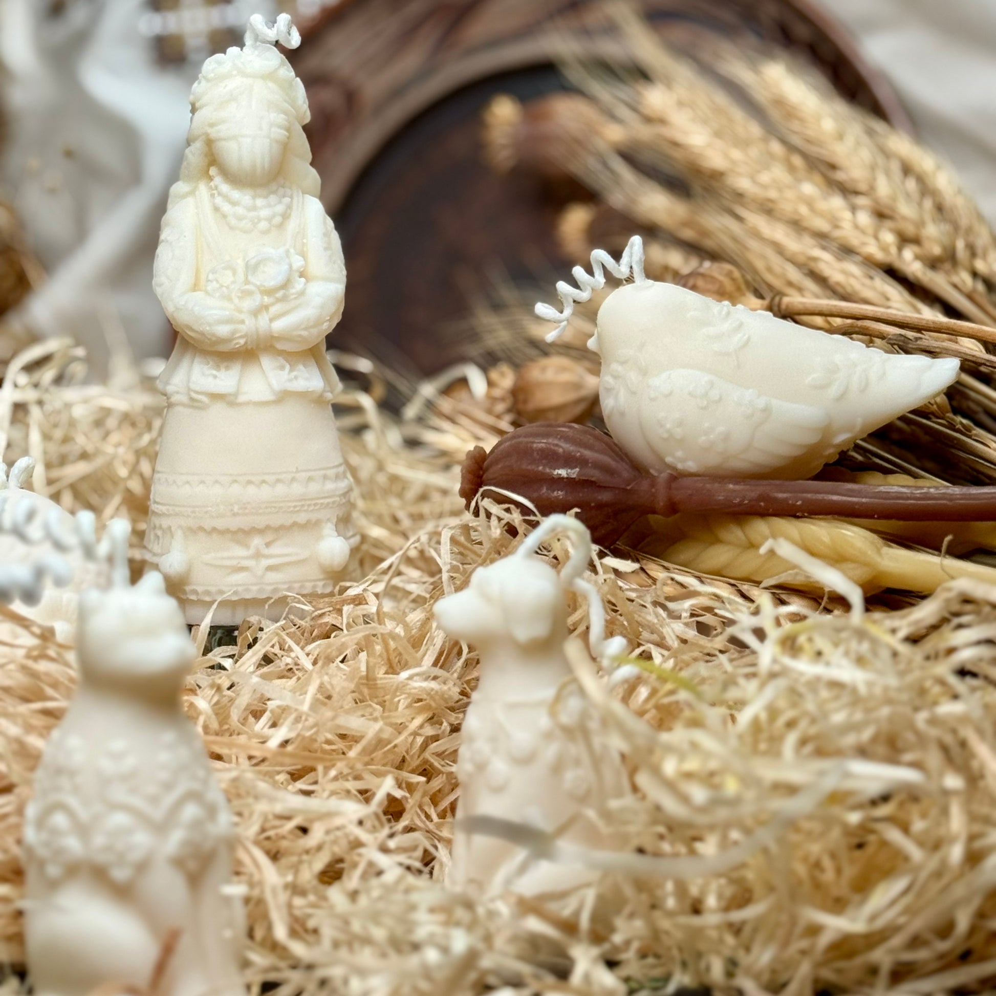 A rustic flat lay display showing the botanical bird, dog, and cat candles nestled in decorative shredded paper alongside a traditional wax figurine and dried poppy pods.