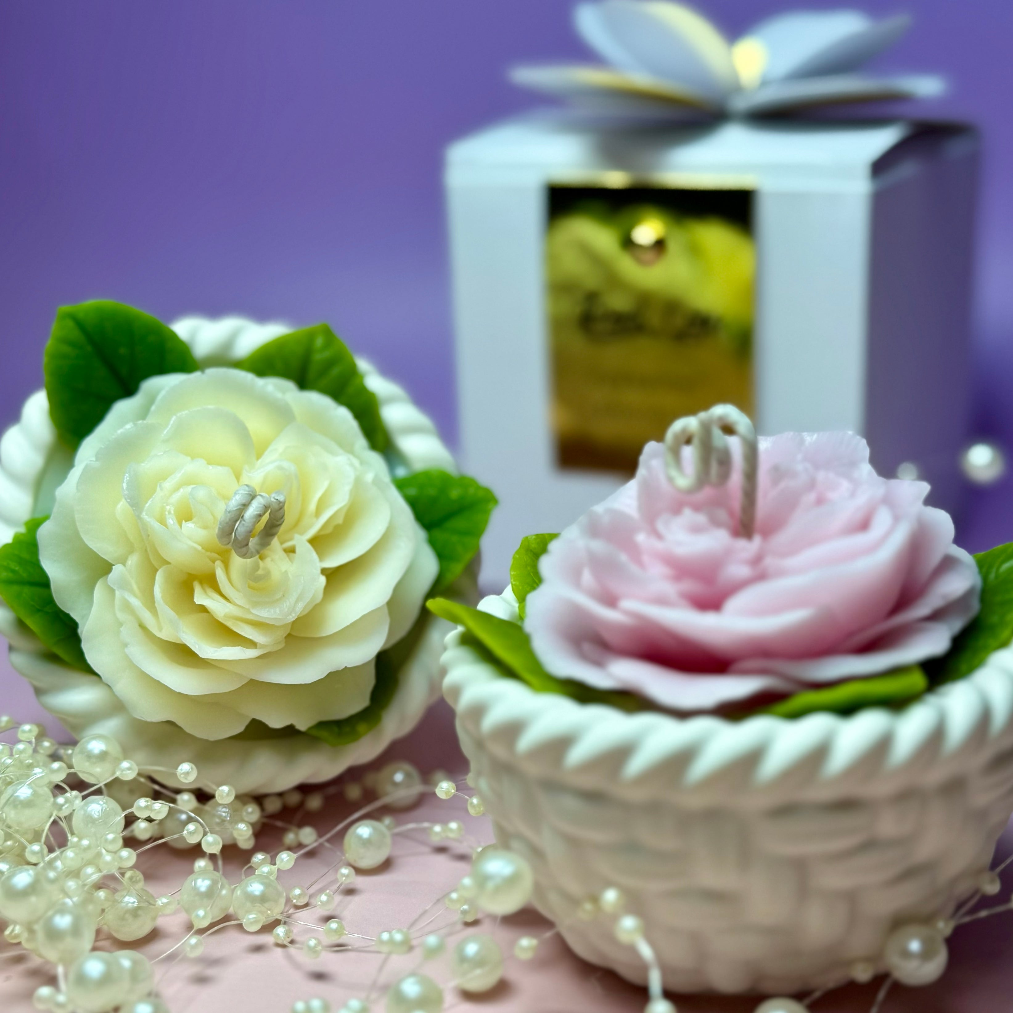 Side-by-side view of two artisan rose candles in woven baskets, showing one in creamy ivory white and one in soft pink with green leaves against a pastel background.